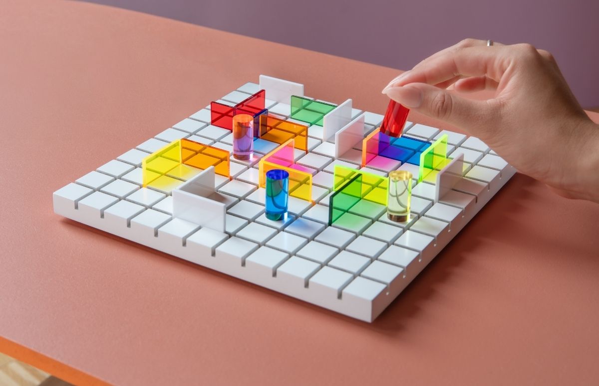 Eine Hand platziert einen roten, durchscheinenden Zylinder auf einem weißen Spielbrett mit Gittermuster. Das Brett ist mit vielen farbigen, transparenten Wänden und Zylindern sowie einigen weißen, undurchsichtigen Wänden wie ein Labyrinth gefüllt.