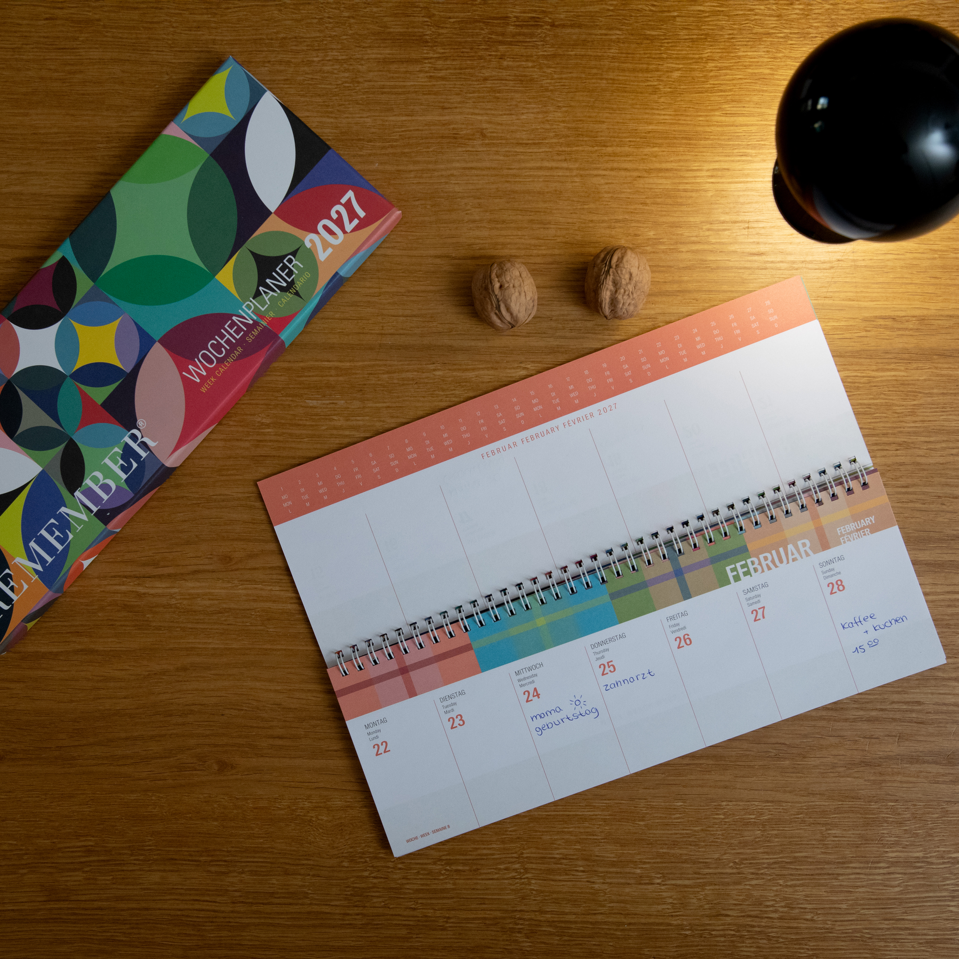 Calendrier hebdomadaire ouvert de février 2027 sur un bureau en bois, avec notes manuscrites comme « Anniversaire de maman » et « Café + gâteau ». À gauche, l’emballage « Agenda hebdomadaire 2027 ». Deux noix et une lampe noire visibles.