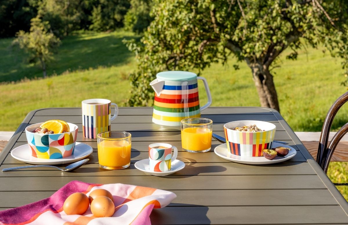 Bunter Frühstückstisch im Freien mit Müsli, Orangensaft und Kaffee. Farbige Schalen, Tassen und gestreifte Teekanne auf dunkelgrauer Platte. Drei Eier auf rosa-orangenem Tuch davor. Im Hintergrund sonnige, grüne Wiese mit Bäumen.