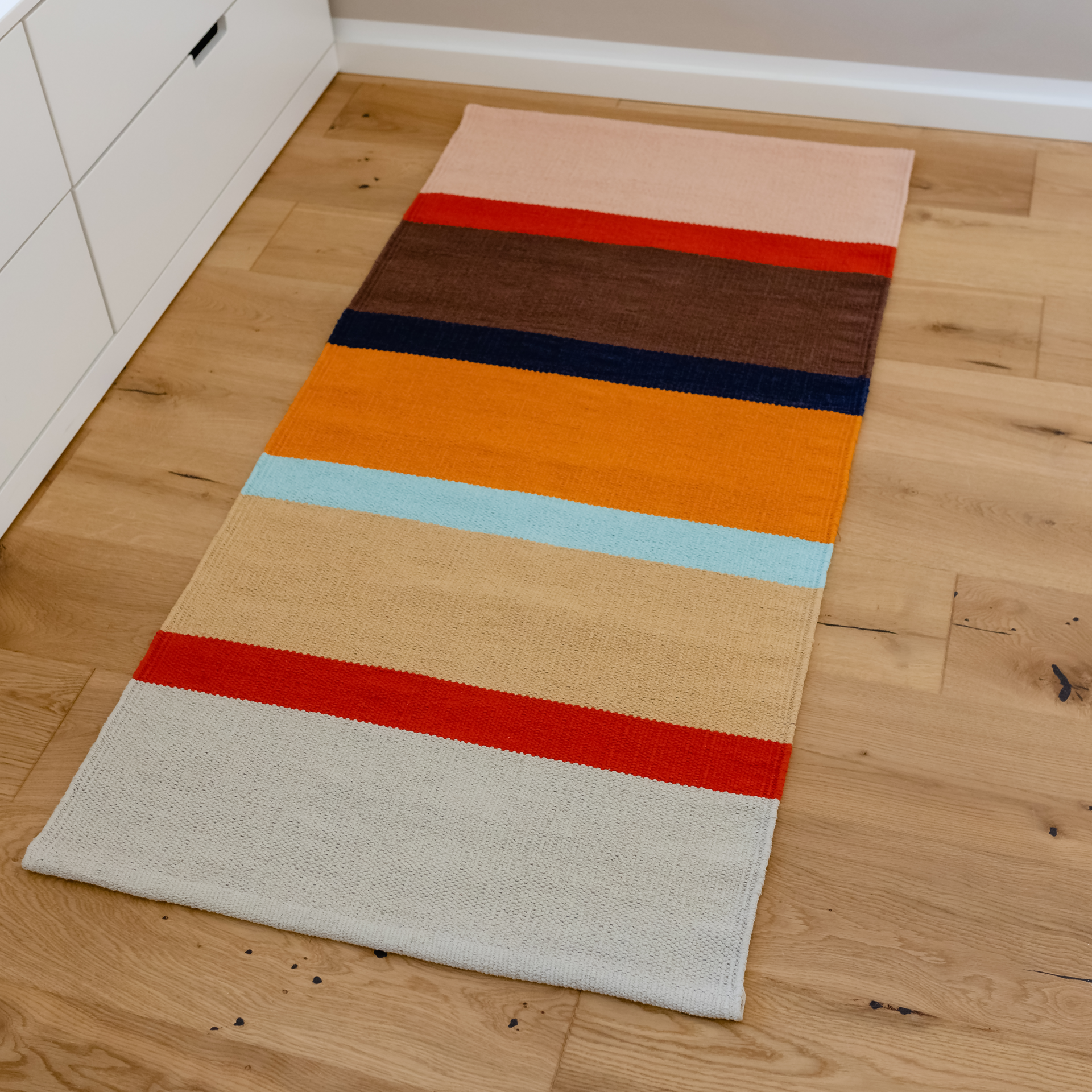 A short-pile carpet runner 'Zaza' lies on a light-coloured wooden floor. The rug has wide stripes in pink, red, brown, dark blue, orange, light blue, beige and grey.