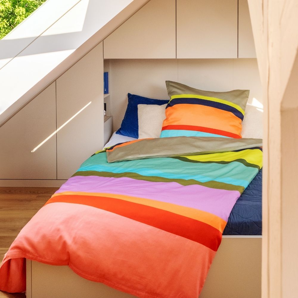 Gemütliches Schlafzimmer unterm Dach mit Bett und bunter Bettwäsche mit breiten horizontalen Streifen in Orange, Rot, Pink, Lila, Blau und Grün. Dahinter weiße Einbauschränke und ein Fenster mit Sonnenlicht und grünem Laub.
