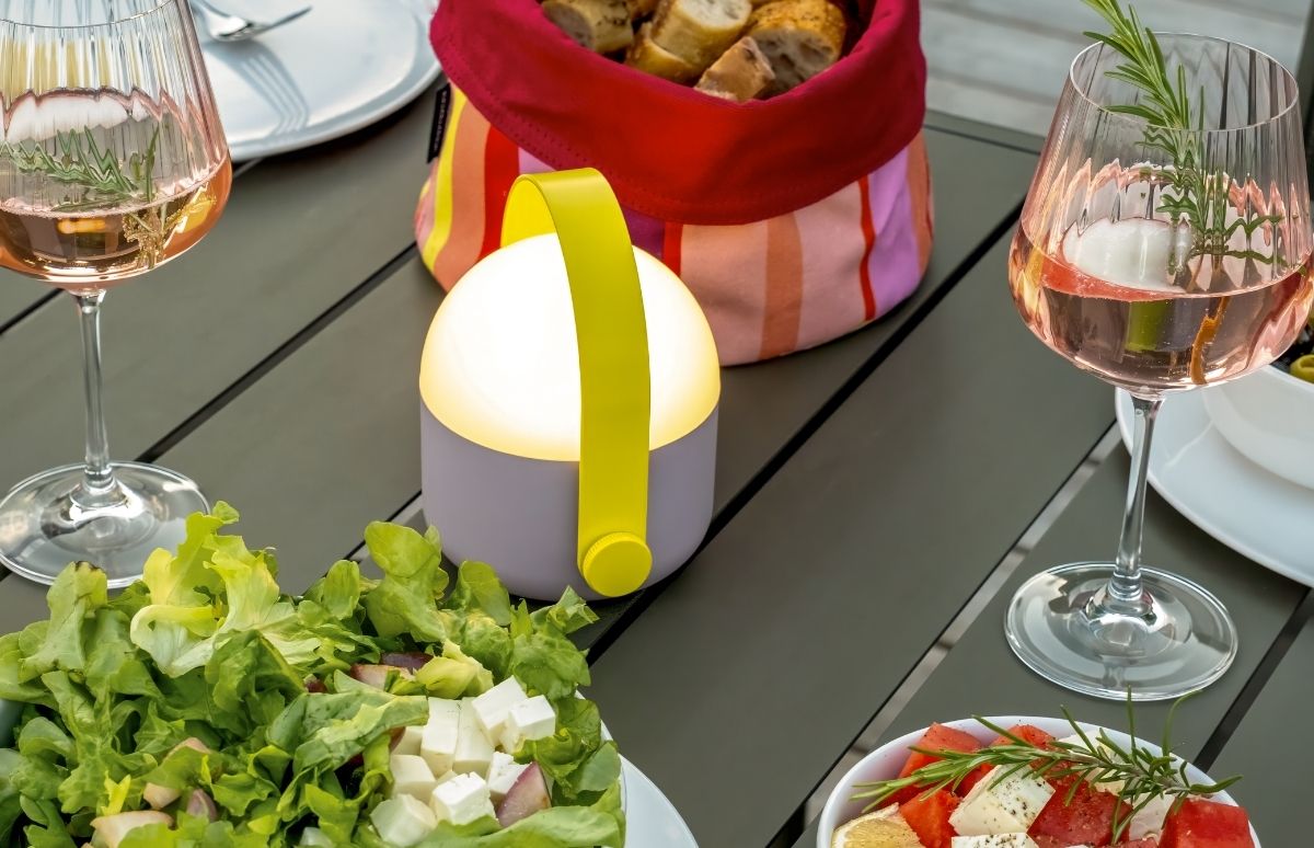 Gedeckter Outdoor-Esstisch im Abendlicht mit warm leuchtender, tragbarer Laterne (gelber Henkel) in der Mitte. Zwei Gläser Roséwein mit Rosmarinzweig, Brotkorb mit Baguette, grüner Salat mit Feta und Wassermelonen-Feta-Salat.