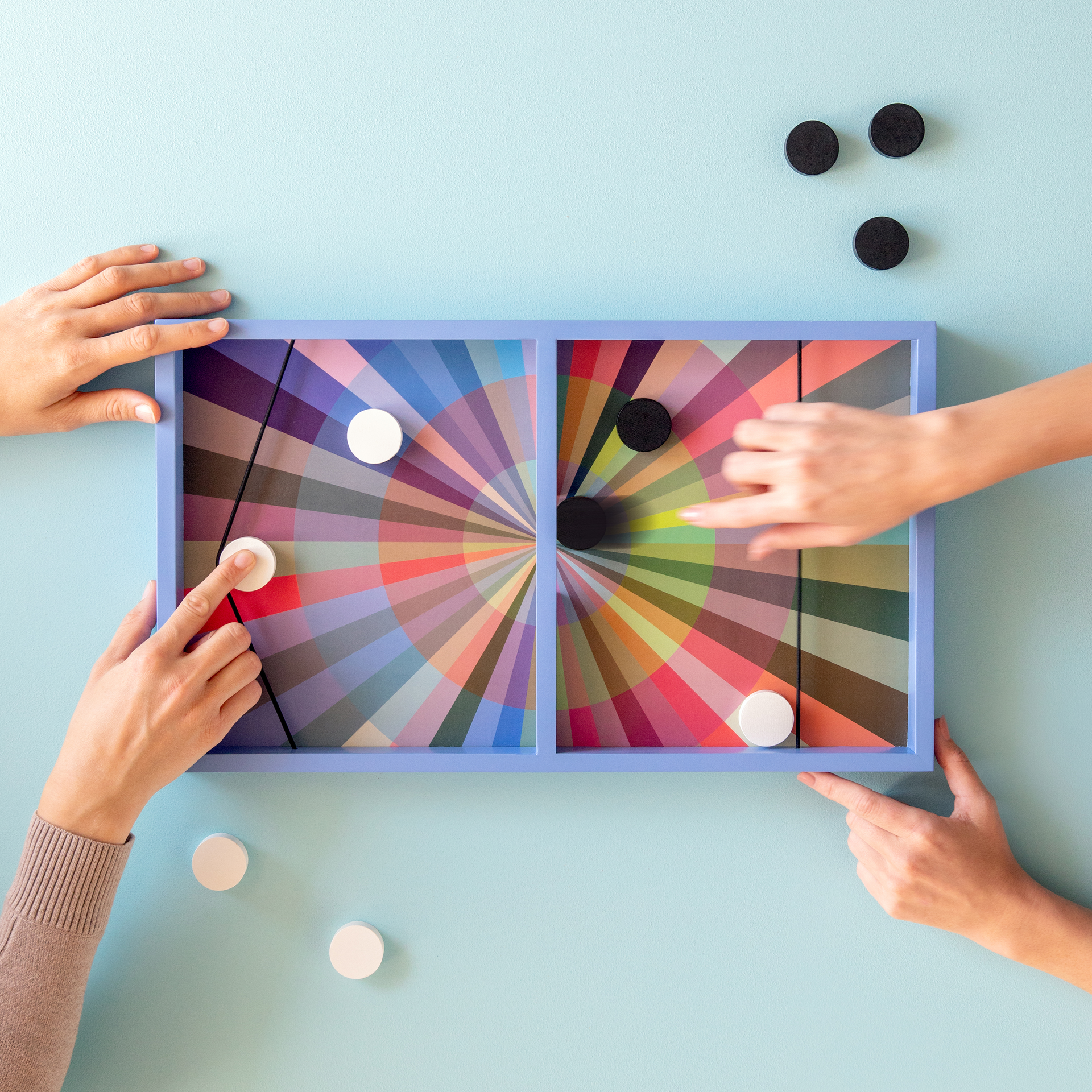Rechteckiges blaues Holzspielbrett für das Geschicklichkeitsspiel „Puck“. Oberfläche in zwei Hälften mit lebhaftem radialem Farbmuster. Schwarze und weiße runde Spielsteine auf und neben dem Brett. Gummibänder als Abschussvorrichtung.