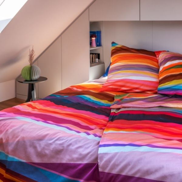 Gemütliches Dachgeschoss-Schlafzimmer: Bett mit lebhaft gestreifter Bettwäsche in Rot-, Orange-, Pink-, Lila- und Blautönen. Links ein kleiner Beistelltisch mit moderner grüner Dekovase. Dahinter weiße Einbauschränke mit Regal.