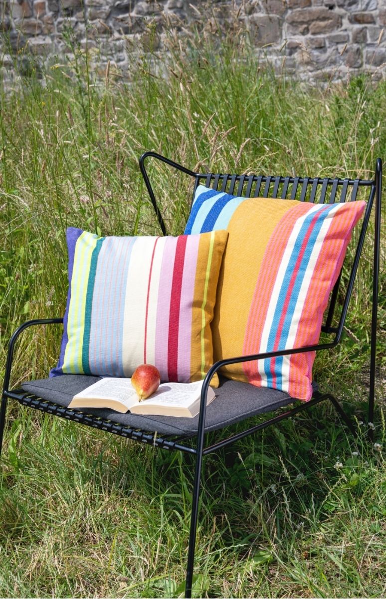 Ein schwarzer Gartenstuhl steht im hohen grünen Gras vor einer Steinmauer. Auf ihm liegen zwei bunte, gestreifte Kissen in leuchtenden Farben. Darauf liegt ein aufgeschlagenes Buch mit einer roten Birne.