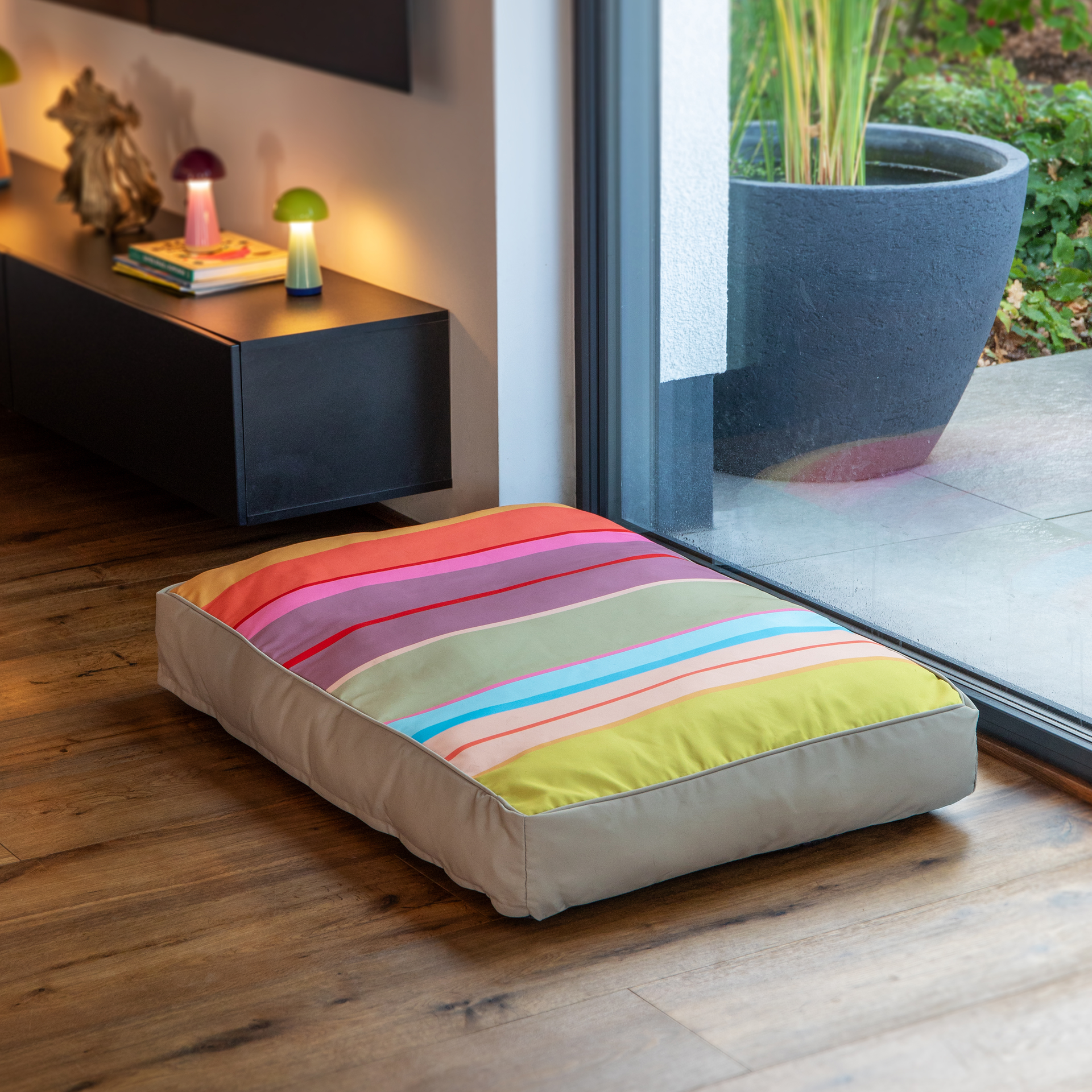 Großes, farbenfrohes Hundekissen mit horizontalen Streifen in Rot, Orange, Pink, Violett, Grün und Blau, umrandet von hellem Beige. Es liegt auf Holzboden vor einer großen Glastür mit Blick in den Garten.
