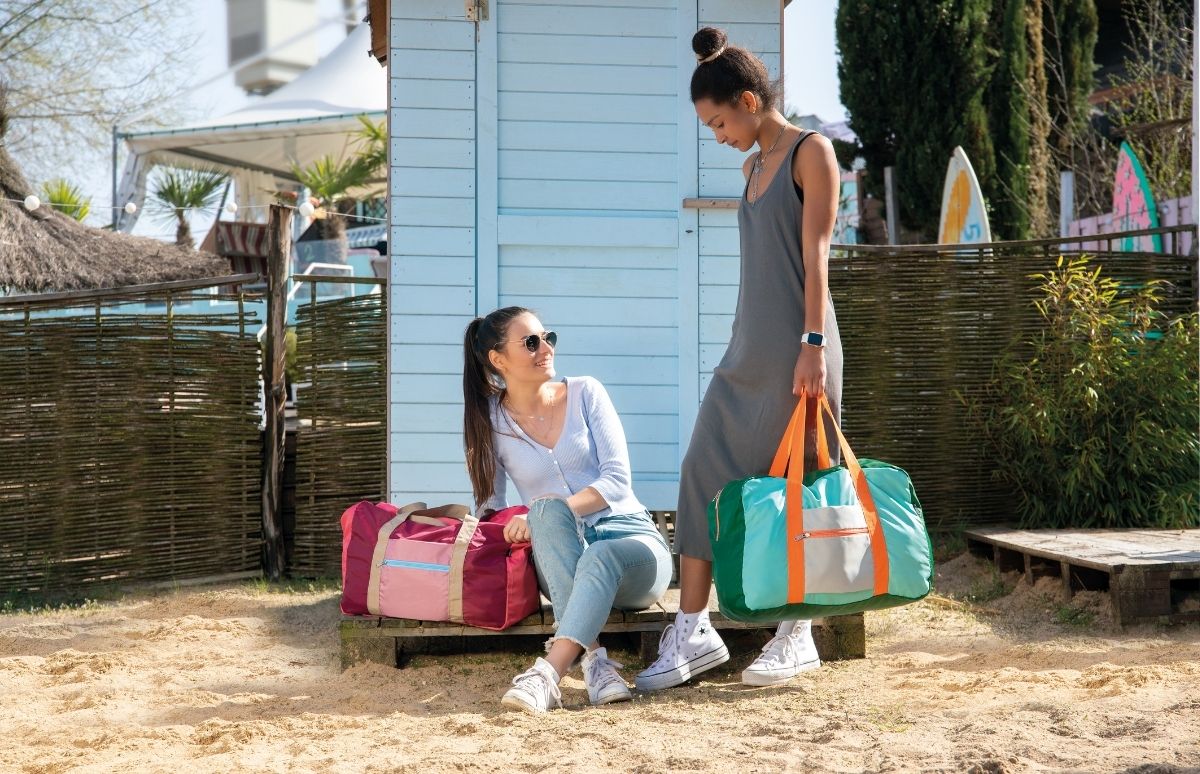 Zwei Frauen am Strand mit bunten Reisetaschen. Eine sitzt auf einer Holzbank, trägt Sonnenbrille und Jeans und lächelt zu ihrer stehenden Freundin mit grüner Tasche. Im Hintergrund: hellblaue Strandhütte und Bambuszaun.