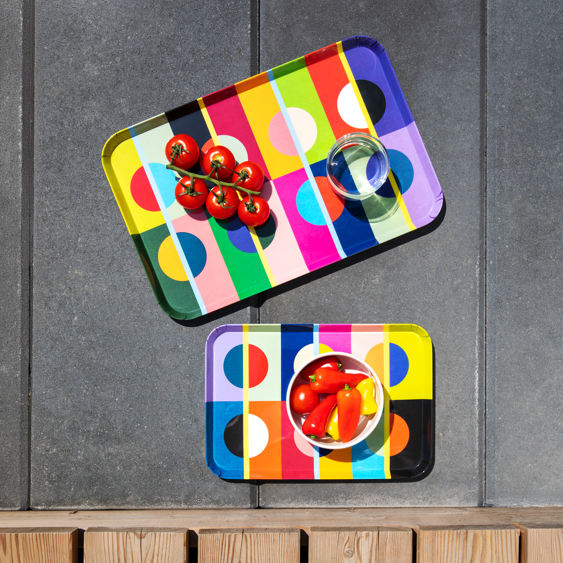 Zwei rechteckige Mini-Tabletts im 'Orb'-Design auf grauem Steinboden. Oben ein größeres Tablett mit roten Kirschtomaten und einem Glas Wasser. Unten eine weiße Schale mit roten und gelben Paprika sowie einer Tomate.