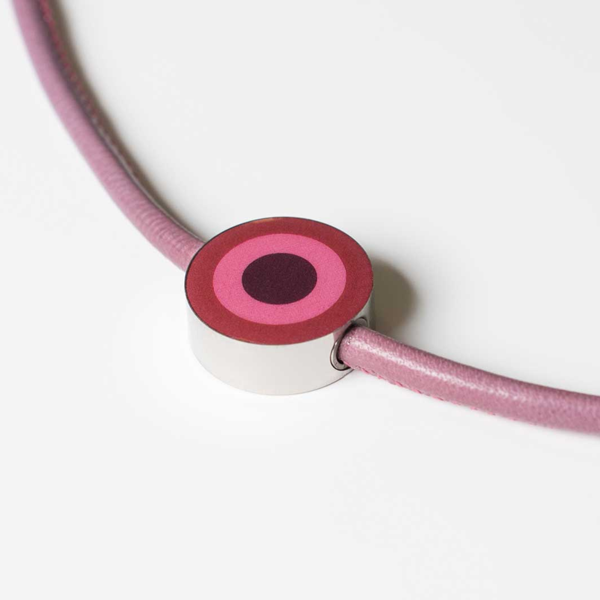 Gros plan sur le pendentif 13, un ruban lilas est maintenu par un pendentif rond argenté. Sur le pendentif, on peut voir des cercles concentriques roses et bordeaux.
