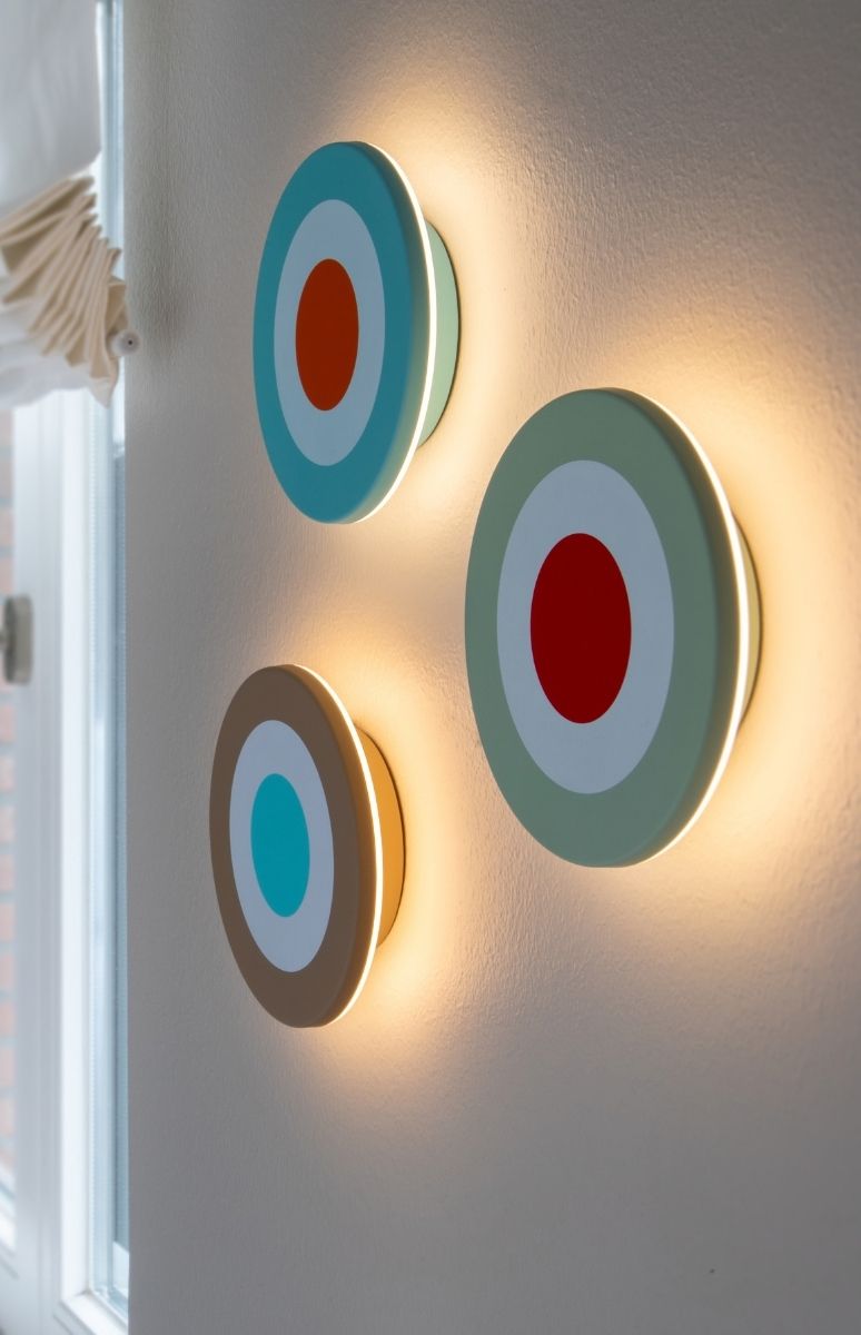 Drei runde Wandleuchten mit markanten Zielscheibenmustern an einer hellen Wand erzeugen warmes Licht. Oben Türkis/Weiß/Orange, mittig Blassgrün/Weiß/Rot, unten Braun/Weiß/Türkis.