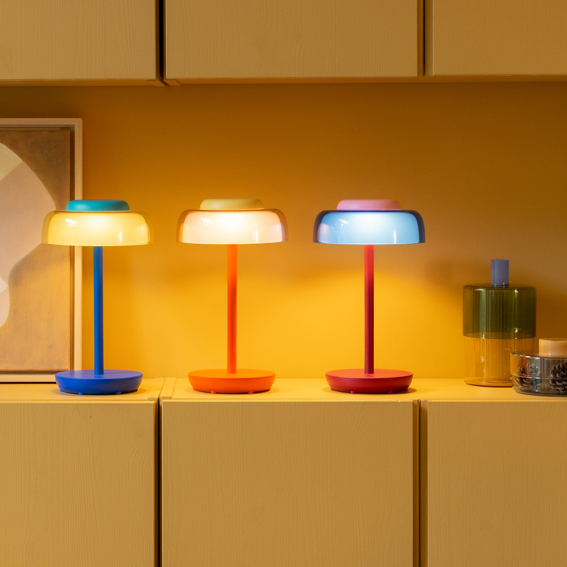 Drie Juno-tafellampen in verschillende kleuren staan op een plank: blauw met een gele kap en een turquoise kapje, feloranje met een geel accent en rood met een blauwe kap en een roze kapje. Moderne draadloze LED-lampen tegen een honinggele muur.