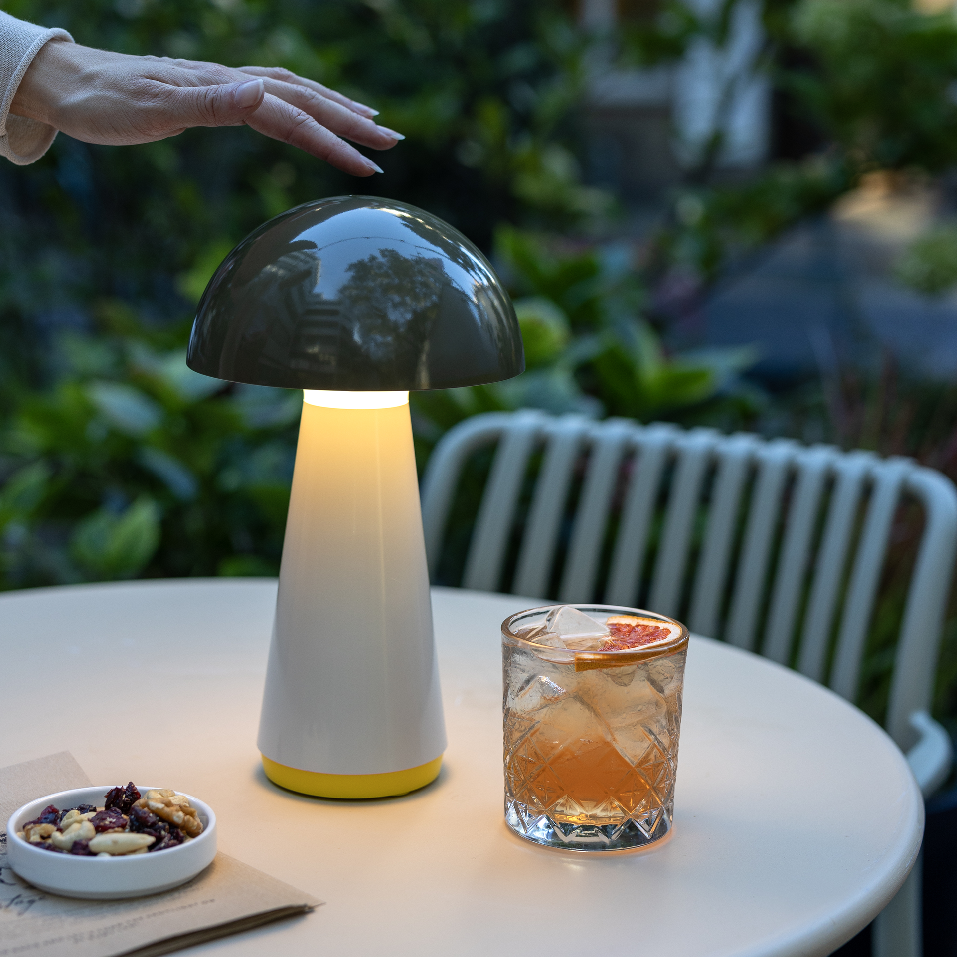 Una lámpara de mesa LED de color oliva con forma de seta está encendida sobre una mesa. Una mano toca la pantalla. Junto a ella, un cóctel y un cuenco con frutos secos.