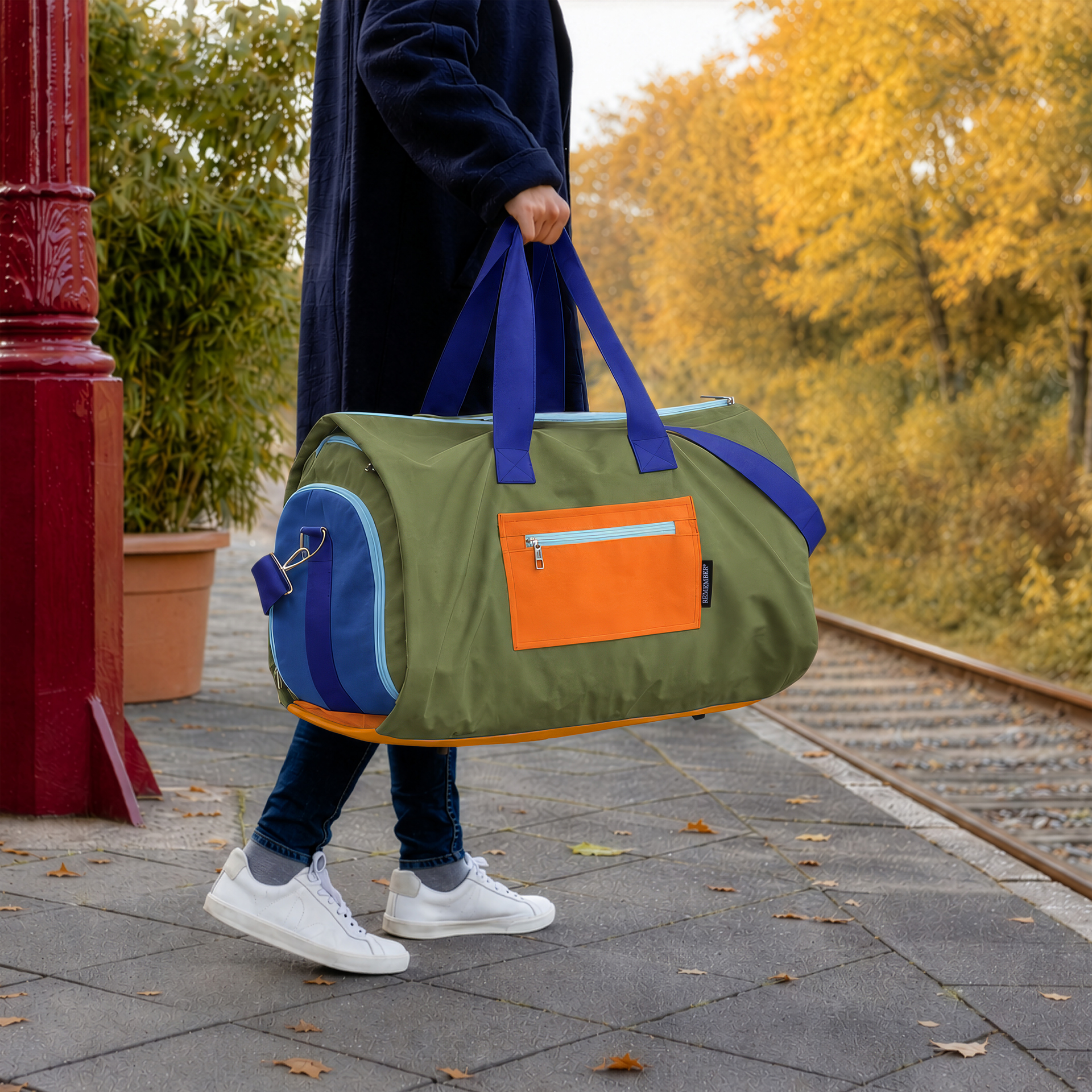 Olivegrüne Reisetasche Lennard, schräg gezeigt. Mit blauen Tragegriffen, Schultergurt mit orangefarbenem Polster und dunkelblauen Seitenfächern mit hellblauen Reißverschlüssen. Wasserabweisend, ideal für unterwegs.