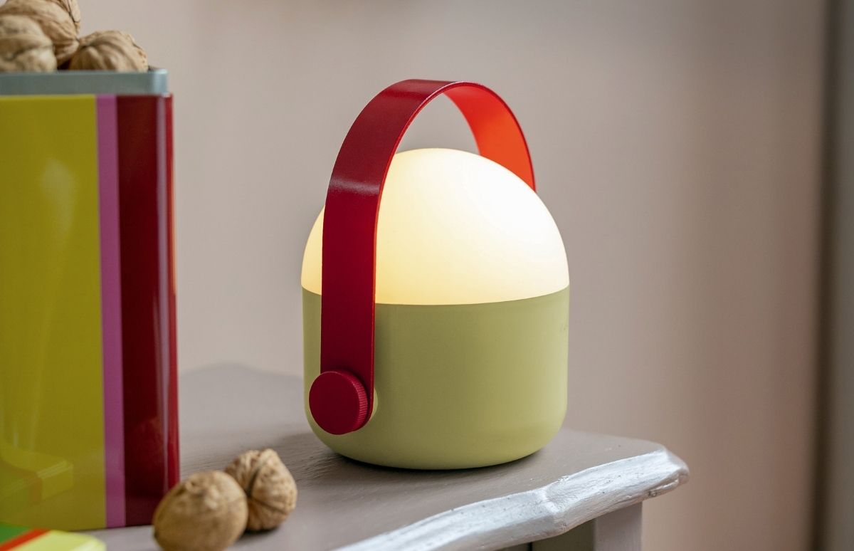 Moderne, tragbare Lampe mit hellgrünem Sockel und leuchtendem, weißem Kugellicht steht auf einem grau gestrichenen Holztisch. Darüber wölbt sich ein auffälliger roter Griff. Links: bunter Behälter und Walnüsse.