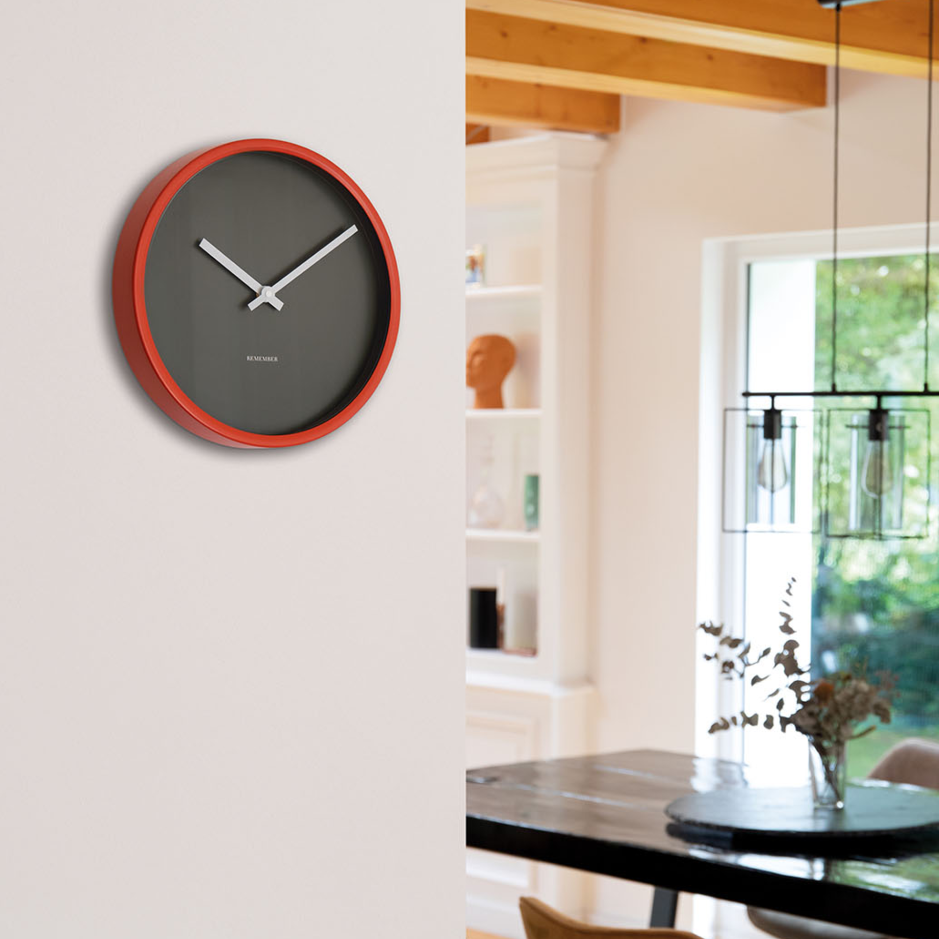 Reloj de pared «Mocca» en una pared blanca. El reloj tiene una carcasa roja, una esfera gris y agujas blancas. Al fondo se ve un comedor con mesa y sillas.