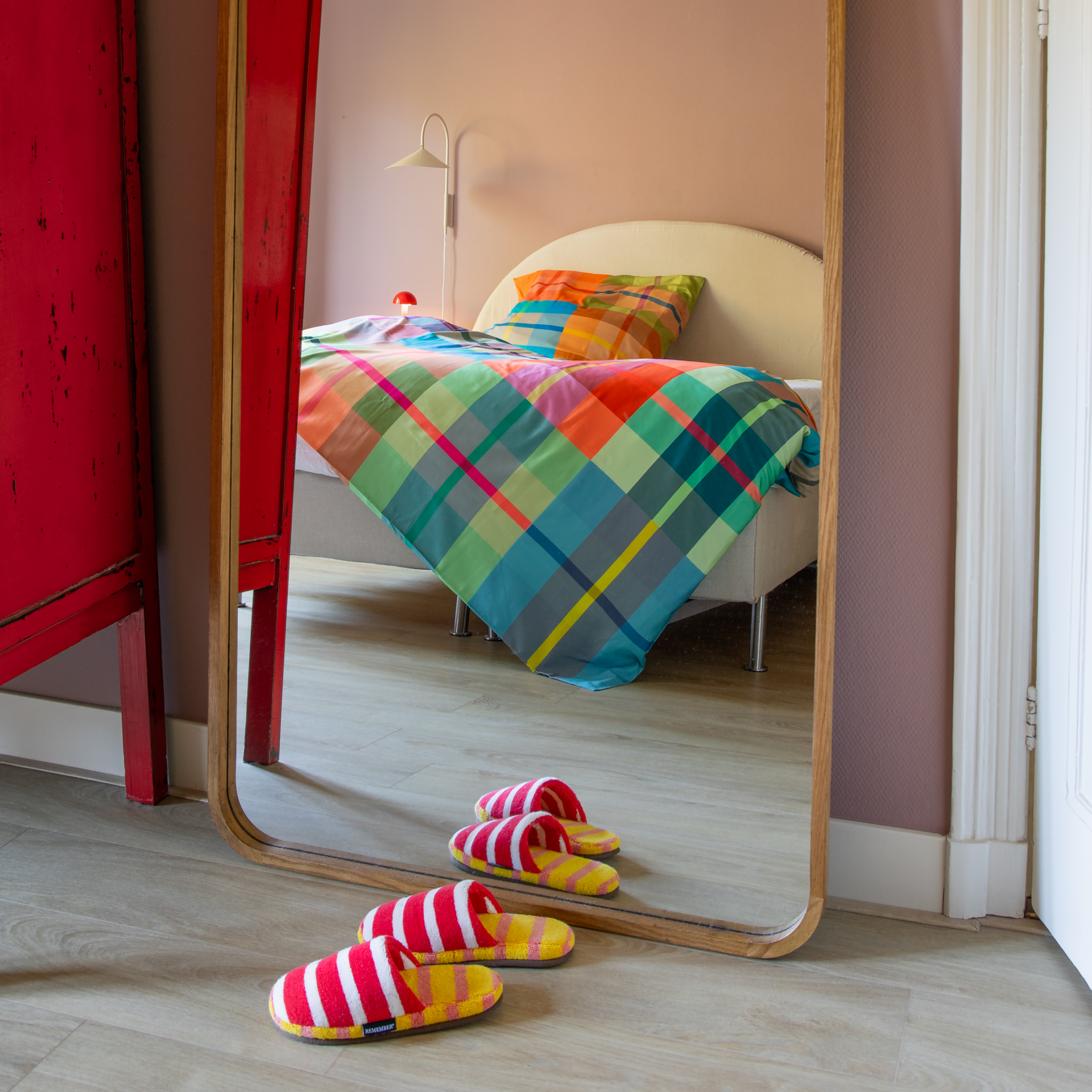 Spiegel zeigt ein Bett mit bunter karierter Bettwäsche. Vorn liegen rot-weiß gestreifte Hausschuhe auf hellem Holzboden. An der rosa Wand: weiße Leselampe und kleine Pilzlampe.