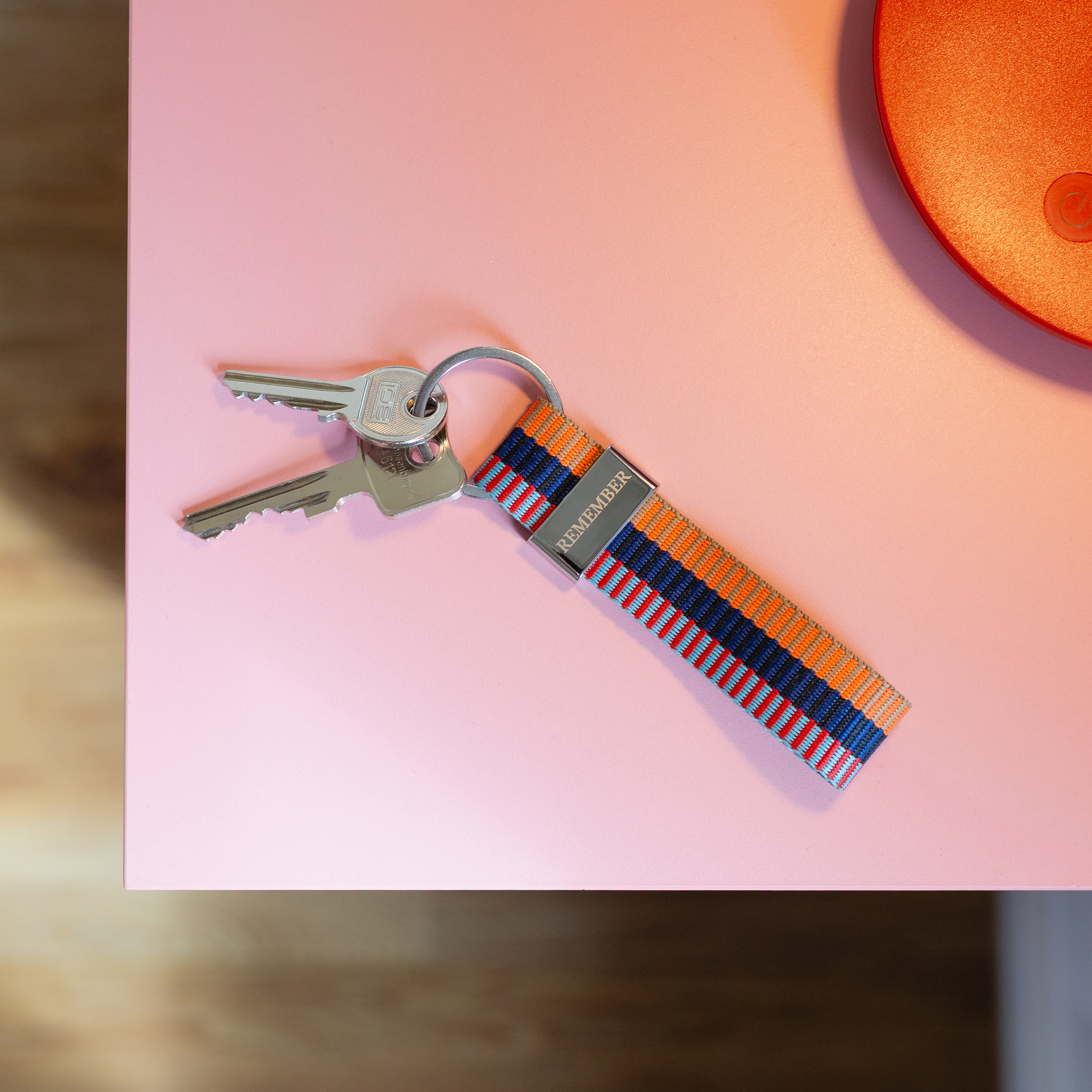 Farbenfrohes Schlüsselband mit blauen, orangefarbenen, roten und hellblauen Streifen, daran zwei silberne Schlüssel. Glänzende Metallplakette mit Gravur „REMEMBER“. Liegt auf rosa Oberfläche, oben rechts Teil eines orangefarbenen Objekts.
