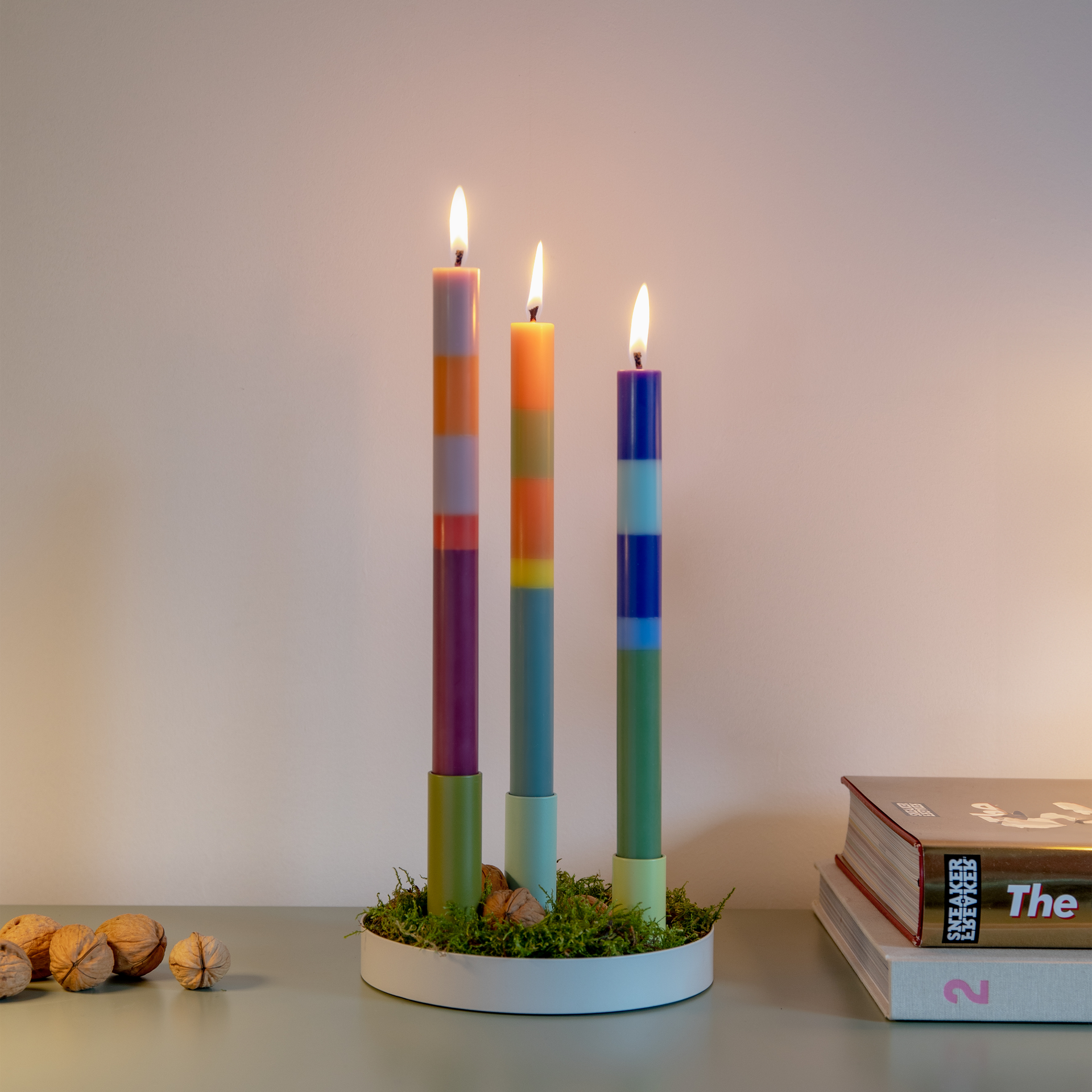 Tres velas de colores encendidas en el portavelas Luna Mini, con musgo y nueces sobre una bandeja blanca. Libros al fondo.