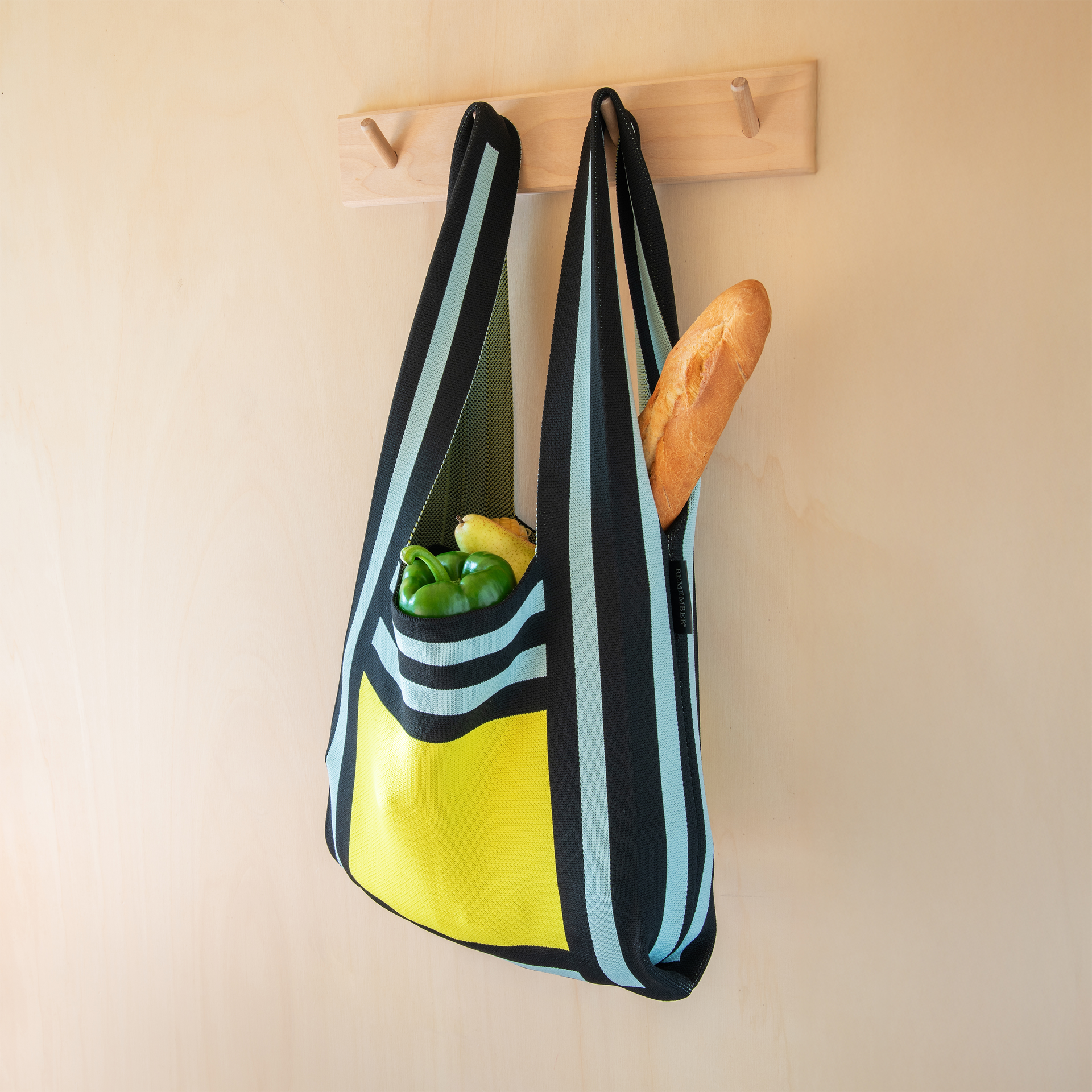 Una bolsa de punto a rayas con alimentos cuelga de un perchero. En la bolsa hay pimientos verdes, una pera y una baguette.