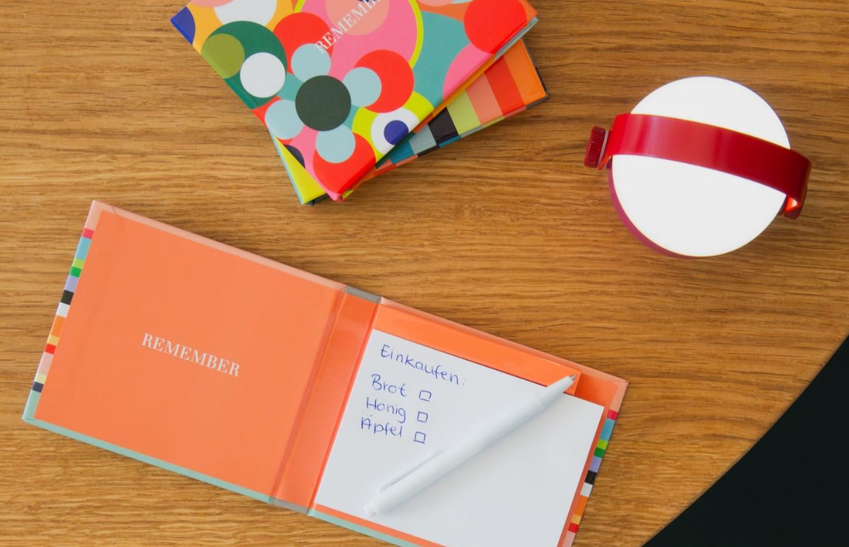 Offenes orangefarbenes Notizbuch auf Holztisch mit handgeschriebener Einkaufsliste ('Brot', 'Honig', 'Apfel') und weißem Stift. Links oben zwei bunte gemusterte Notizbücher. Rechts oben runde weiße Lampe mit rotem Band.