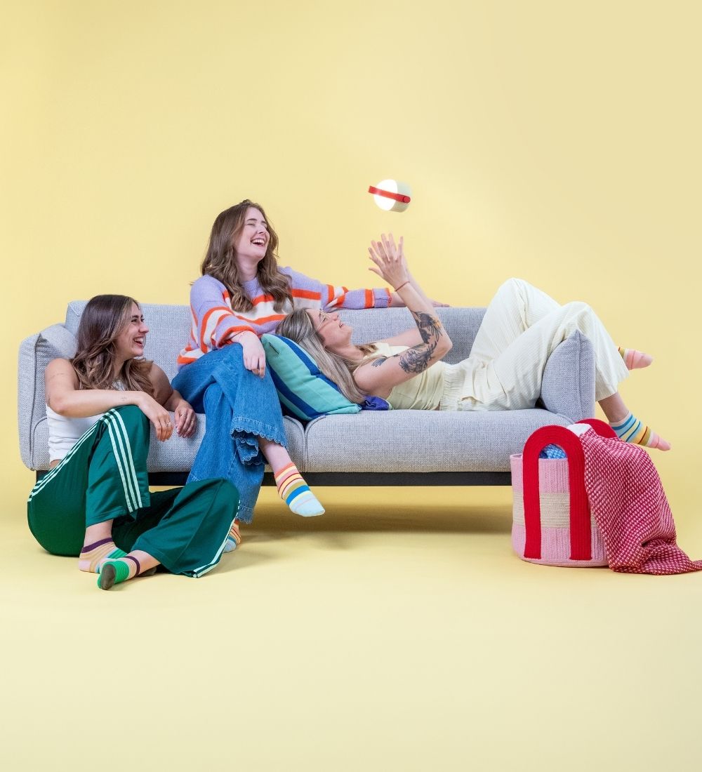 Drei lachende Frauen in bunten Socken entspannen auf und neben einem grauen Sofa vor gelbem Hintergrund: eine sitzt am Boden, eine auf dem Sofa, eine liegt darauf und greift nach einem schwebenden Ball. Daneben steht ein pinker Korb mit roter Decke.