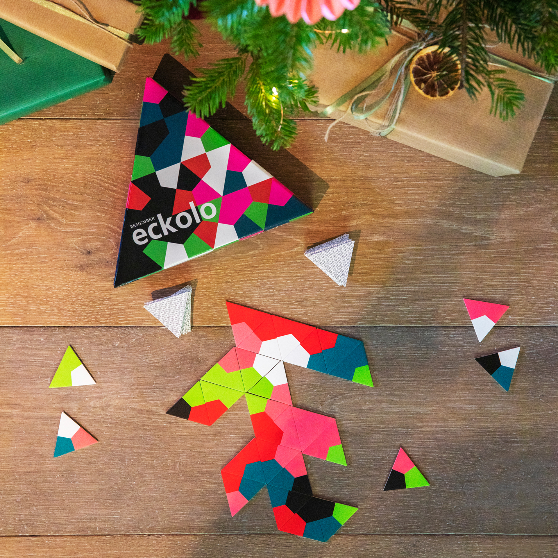 Gioco di Eckolo: carte triangolari, scatola con motivo geometrico e regali su un tavolo di legno sotto un albero di Natale.
