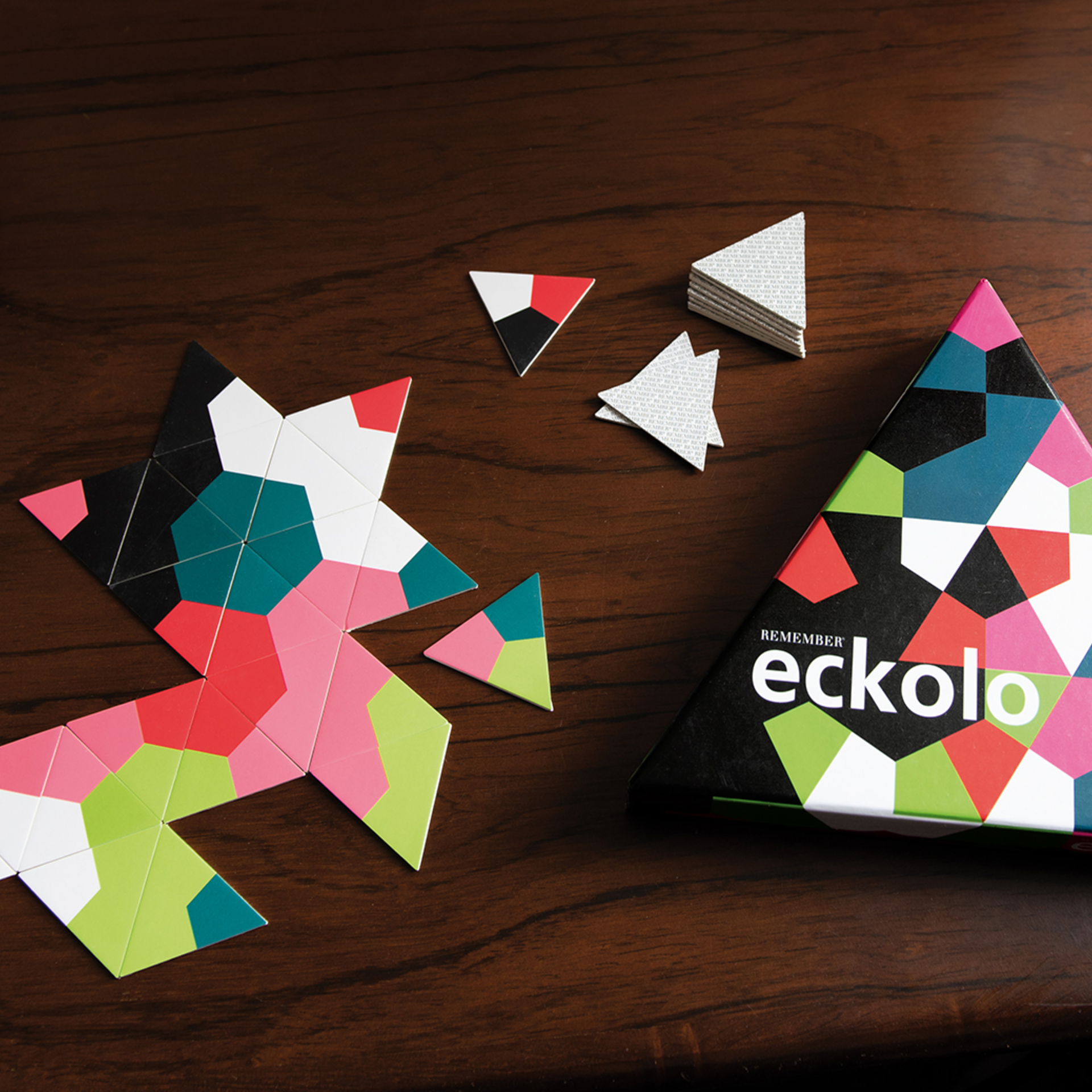 Vista dall'alto: mazzo Eckolo con carte triangolari, alcune disposte a stella. Scatola del gioco a destra. Mazzo di carte e carte sciolte. Colori: Verde, rosa, rosso, nero, bianco, blu-verde.