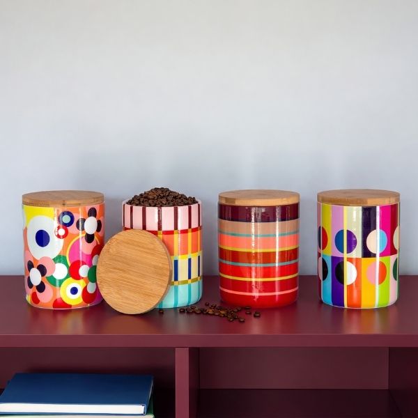 Quatro potes coloridos com tampas de madeira numa prateleira vermelho-escuro. O segundo pote à esquerda está aberto com grãos de café; a tampa está à frente. Padrões modernos com flores, riscas e círculos em amarelo, rosa, azul e vermelho.