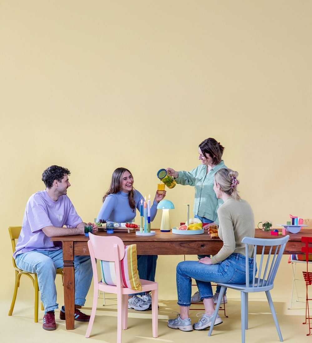 Vier Freunde sitzen lachend an einem Holztisch mit bunten Kerzen, stilvoller Lampe und frischen Früchten. Eine Frau gießt Saft aus einer gelb-grünen Karaffe in ein Glas, während die anderen lächelnd zusehen.