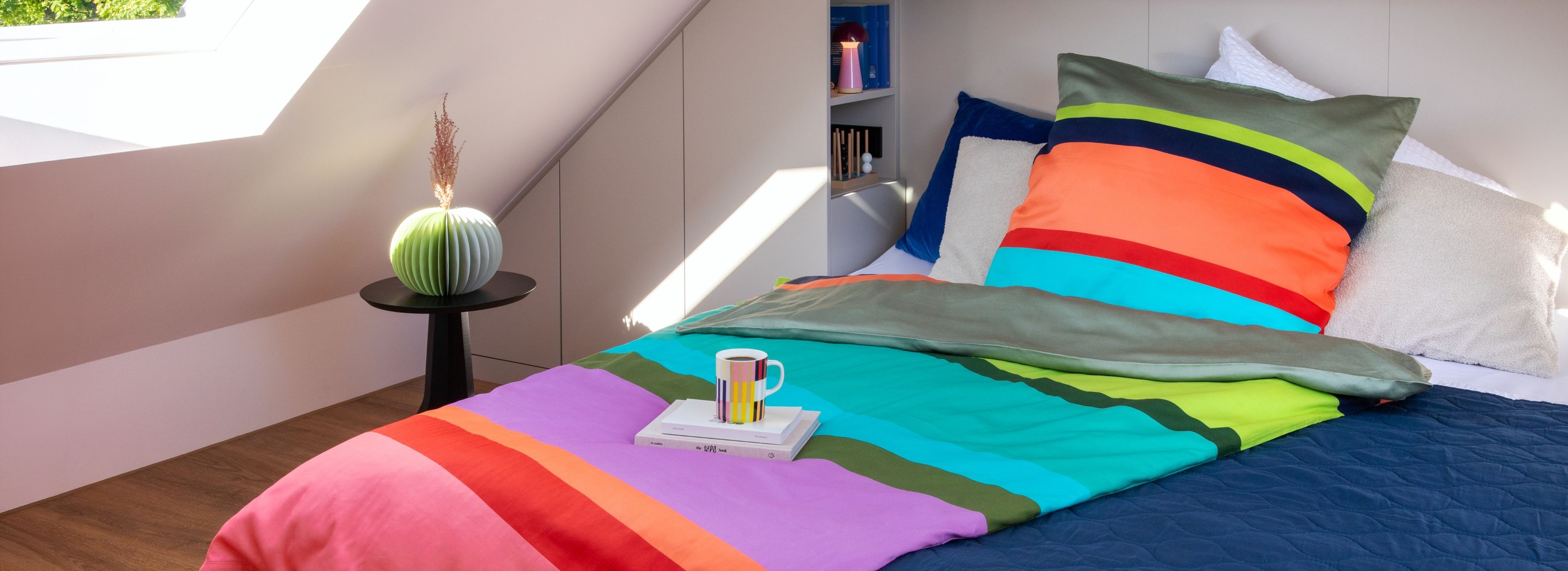 Helles Dachgeschoss-Schlafzimmer. Einzelbett mit bunter, gestreifter Bettwäsche, darauf zwei Bücher und eine Tasse. Daneben kleiner Tisch mit grüner Faltvase und getrockneten Blumen. Regale im Hintergrund. Sonnenlicht durchs Dachfenster.