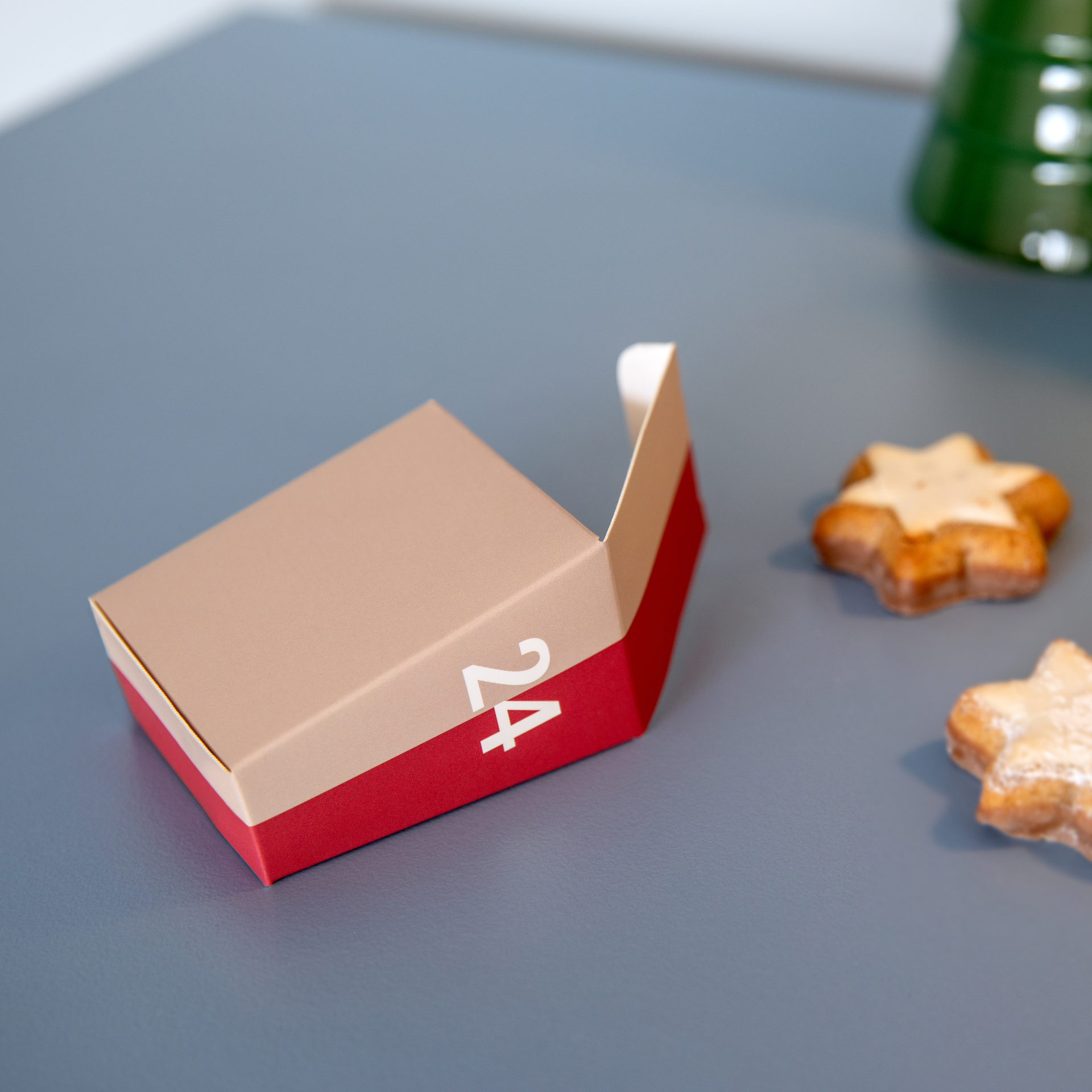 Caja de calendario de Adviento de color rojo y beige abierta, con un «24» blanco sobre un fondo gris. A la derecha hay dos galletas con forma de estrella, ligeramente espolvoreadas con azúcar glas y algo desenfocadas, que realzan el ambiente festivo.