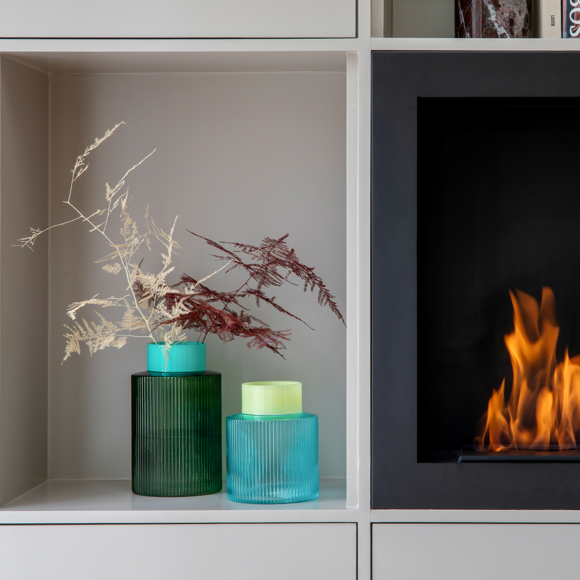 Two glass vases, a blue and a green groove vase, decorated with dried twigs, stand on the shelf next to a fireplace with a burning fire.