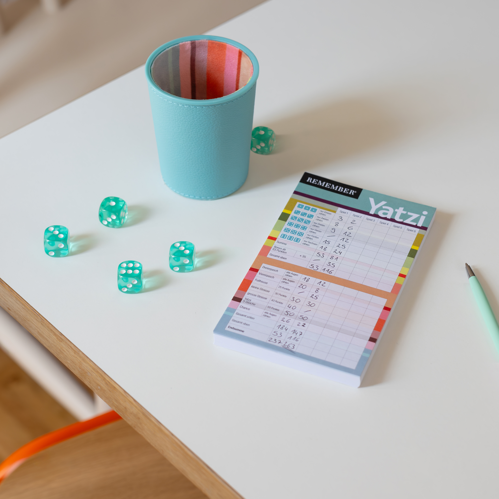 Op een witte tafel staat een turquoise dobbelbeker van kunstleer met een kleurrijk gestreepte binnenvoering. Ernaast liggen vijf doorschijnende groene dobbelstenen. Rechts ligt een Yatzi-notitieblok met spelstanden en een bijpassende pen.