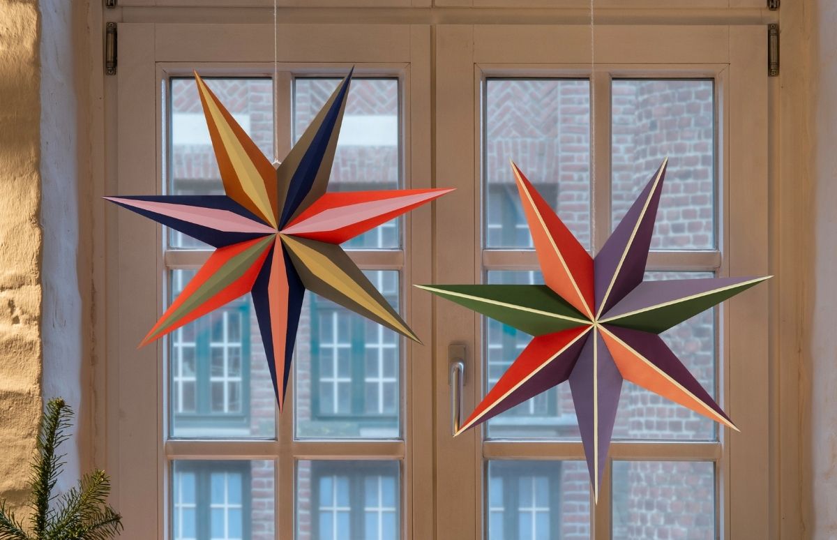 Zwei bunte, geometrische Papiersterne hängen vor einem Fenster. Links Pink, Orange, Blau, Grün; rechts vor allem Rot, Lila, Orange, Grün. Beide sind segmentiert und haben zwölf Spitzen. Im Hintergrund eine Ziegelmauer, unten links ein Tannenast.
