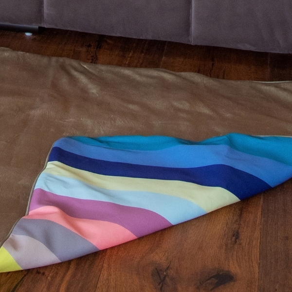 A colourful, striped blanket on a wooden floor. Horizontal stripes in sky blue, dark blue, pastel yellow, pink, purple and grey. Part of it is folded and lies on a light brown carpet. A dark piece of furniture is visible at the top.