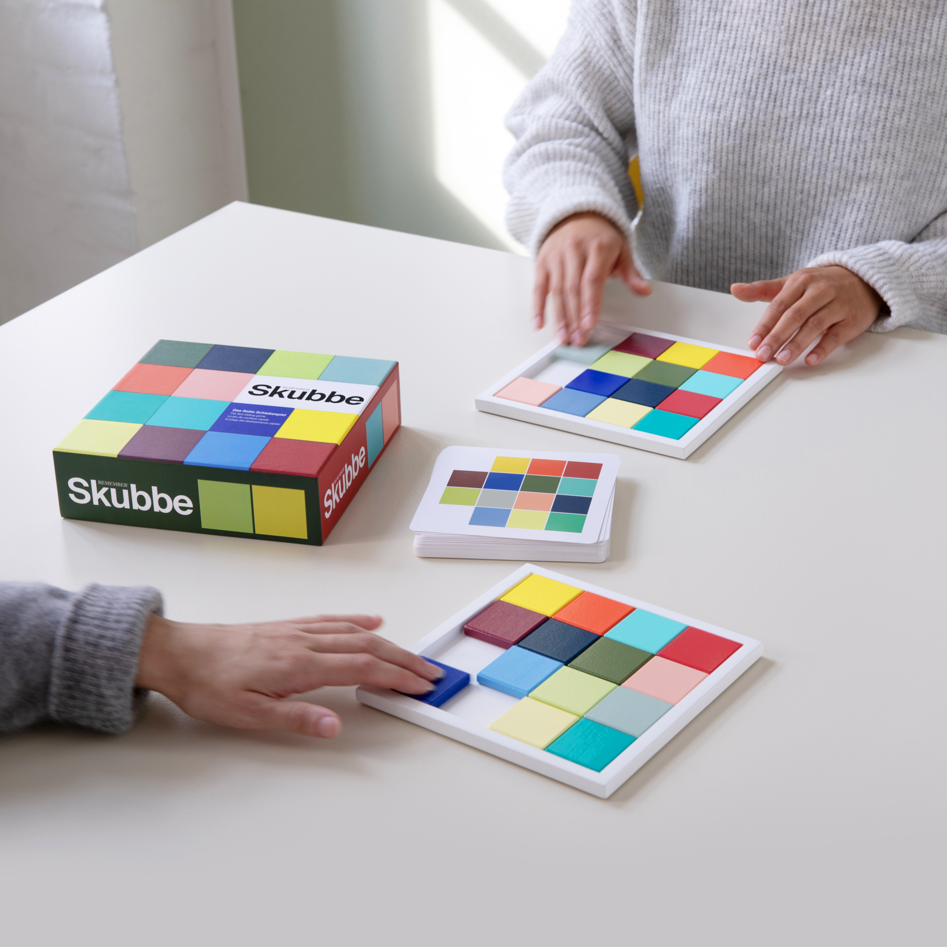 Dos personas juegan a «SKUBBE». Las fichas de madera están dentro de un marco y las cartas con motivos de colores están sobre una mesa. Se ve el packaging del juego «SKUBBE».