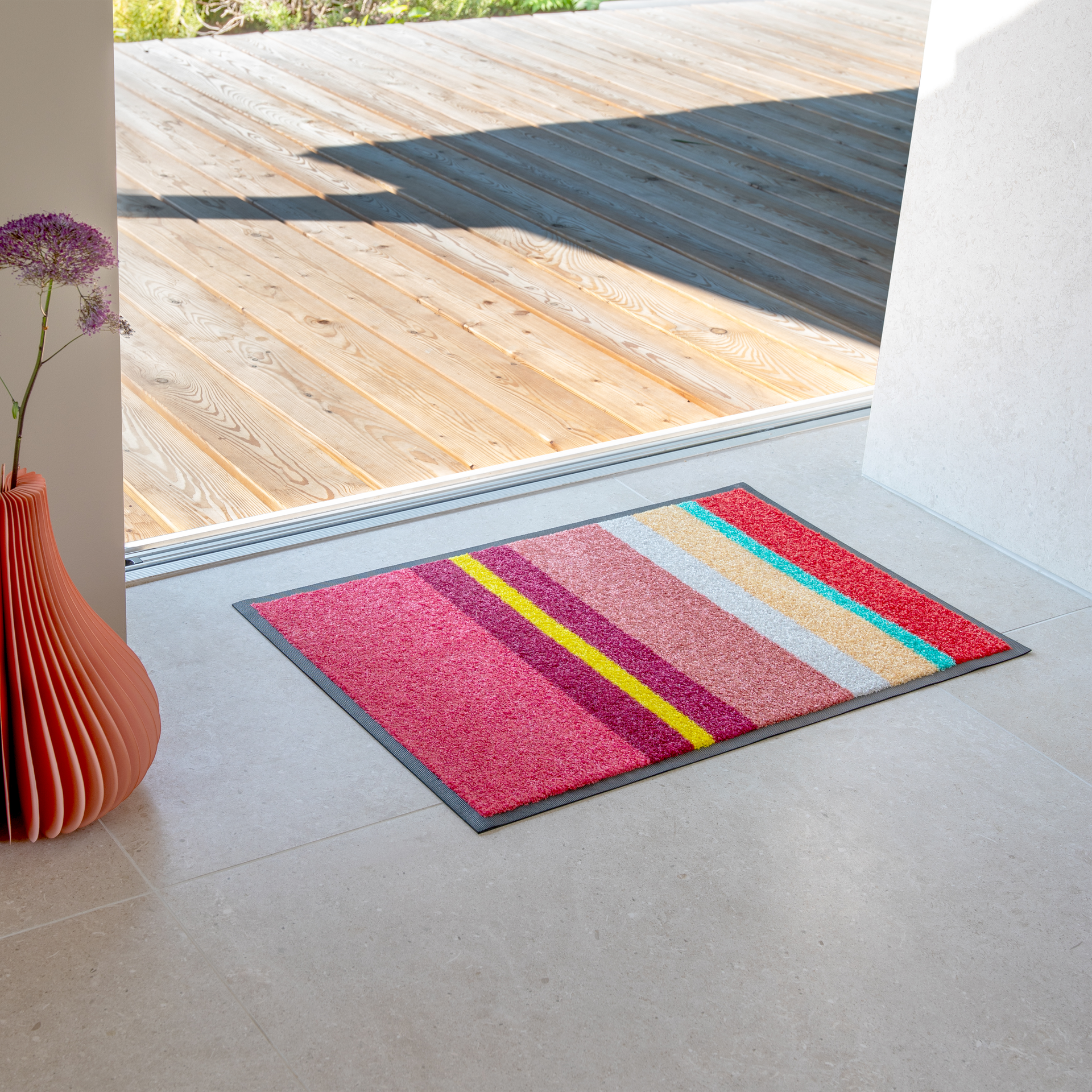 A colourful doormat with diagonal stripes in red, pink, yellow, white and turquoise lies on light grey tiles in front of an open glass door. Behind it, a sunny wooden terrace is visible. To the left stands an orange vase with a purple flower.