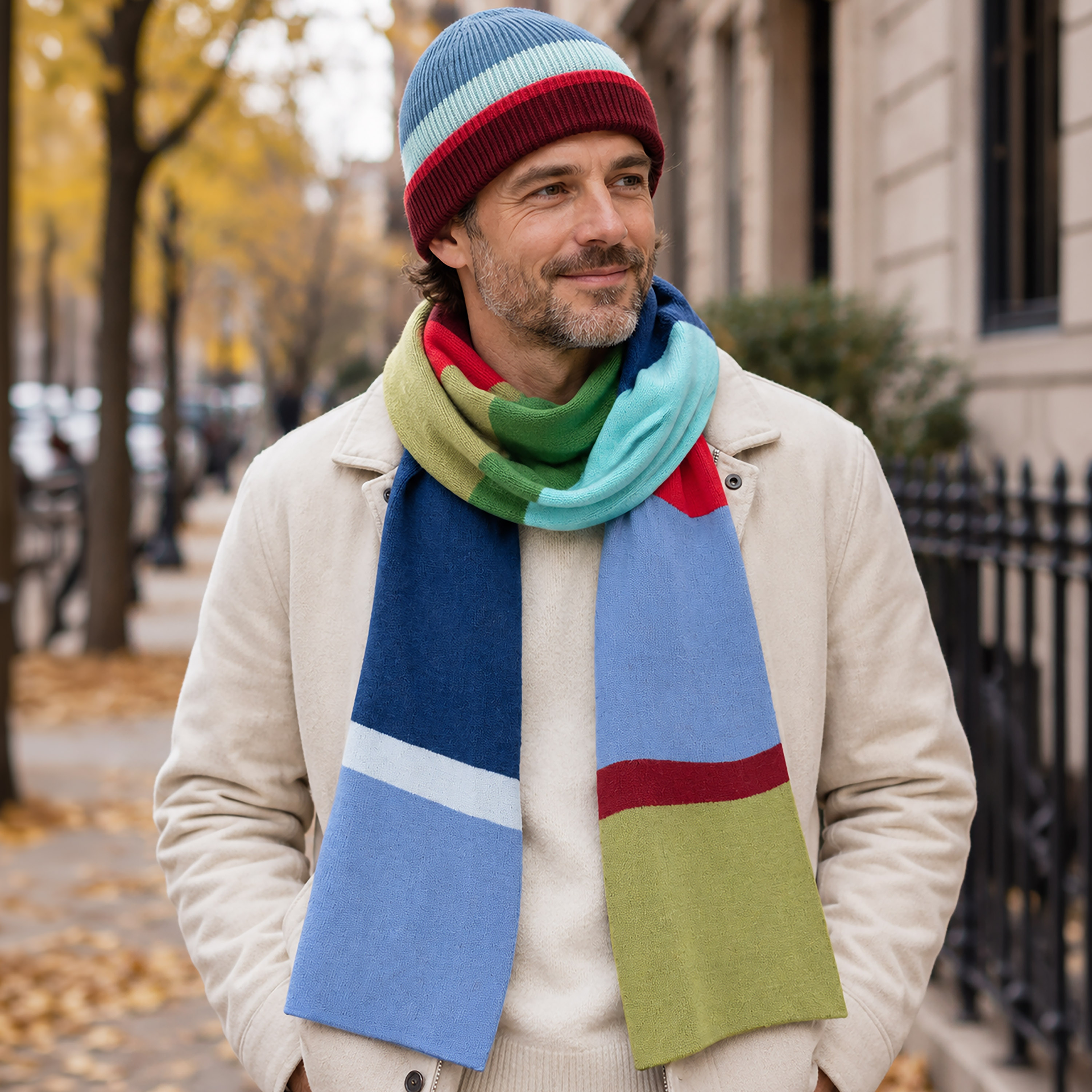 Un hombre sonriente lleva un gorro de lana y cachemira multicolor con rayas horizontales en rojo oscuro, azul claro y azul medio. Lo combina con una bufanda a juego con bloques de color en azul, rojo, verde y azul claro. Está de pie en la acera.