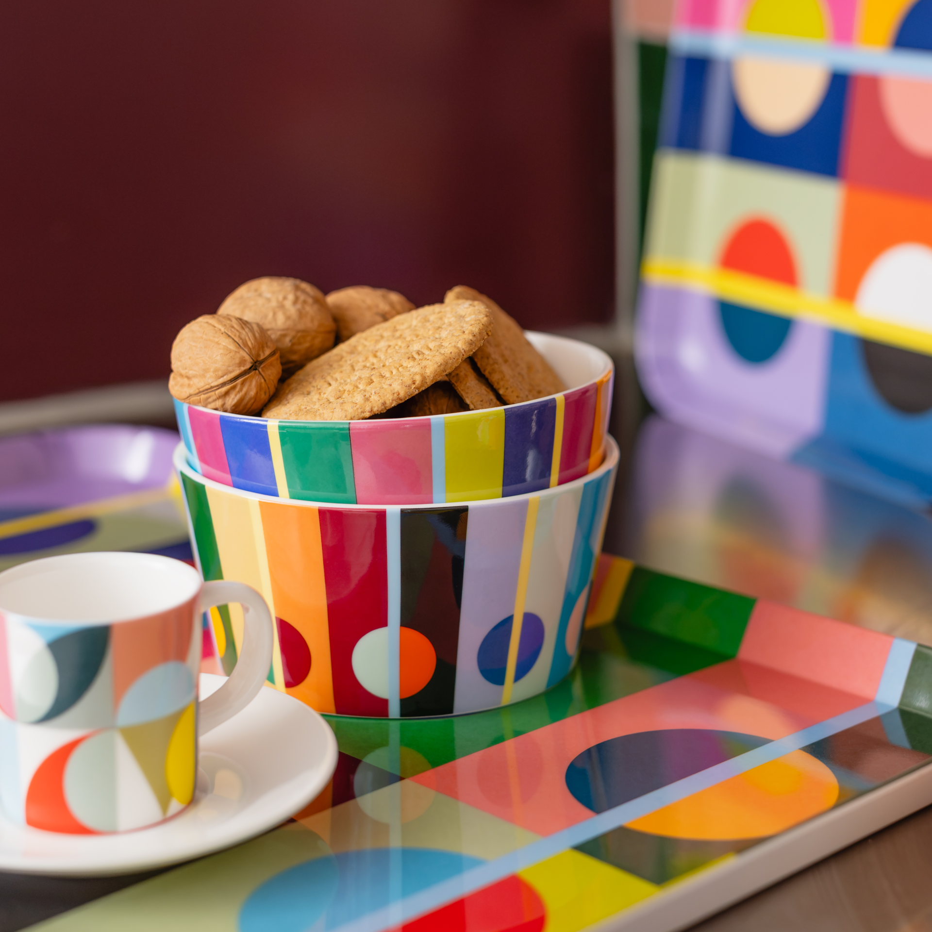 Two colourful, stacked cereal bowls from the Orb collection, filled with walnuts and biscuits. Featuring a vibrant, geometric pattern of colourful stripes and circles. Cup and saucer, and a large tray in the same design.