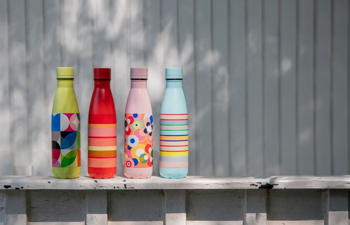 Vier farbenfrohe Trinkflaschen stehen nebeneinander auf einer weiß gestrichenen, leicht verwitterten Holzleiste im Freien: gelb mit geometrischen Formen, rot gestreift, pink mit Blumen, hellblau mit bunten Streifen.