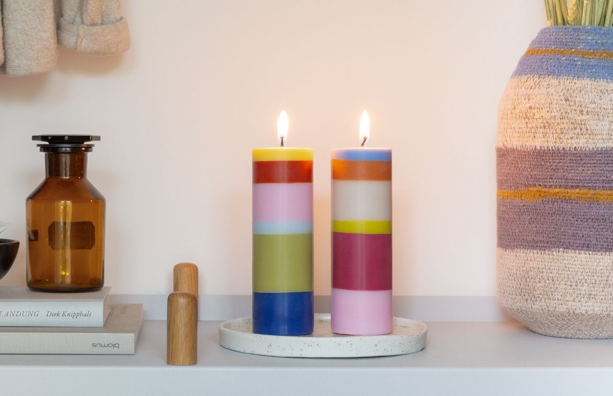 Zwei brennende Kerzen auf einem runden, hellen Tablett. Beide haben bunte Querstreifen in lebhaften Farbkombinationen. Links steht ein braunes Apothekerglas auf Büchern, rechts ein Korb mit getrockneten Gräsern. Wohnlich.