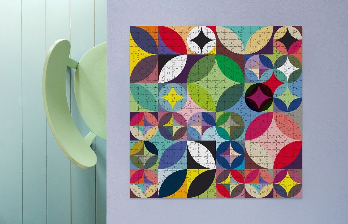 Big puzzle with a colorful abstract pattern of overlapping circles and squares (red, blue, green, yellow, purple, black). A light green chair sits at left before pale blue wood paneling.