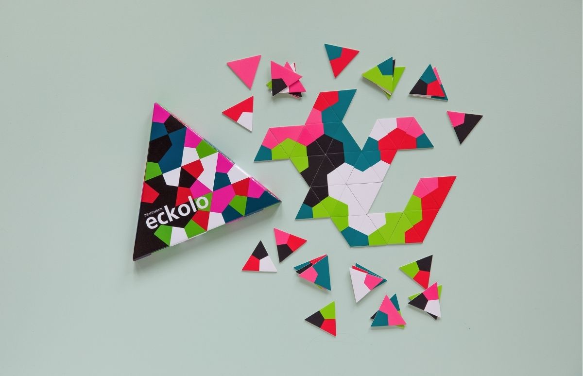 Bunte dreieckige Puzzleteile auf hellgrünem Hintergrund. Links eine dreieckige Schachtel mit "eckolo"-Logo und ähnlichem Muster. In der Mitte bilden viele kleine Dreiecke in Pink, Grün, Türkis, Schwarz und Weiß eine große geometrische Form. Lose Teile.