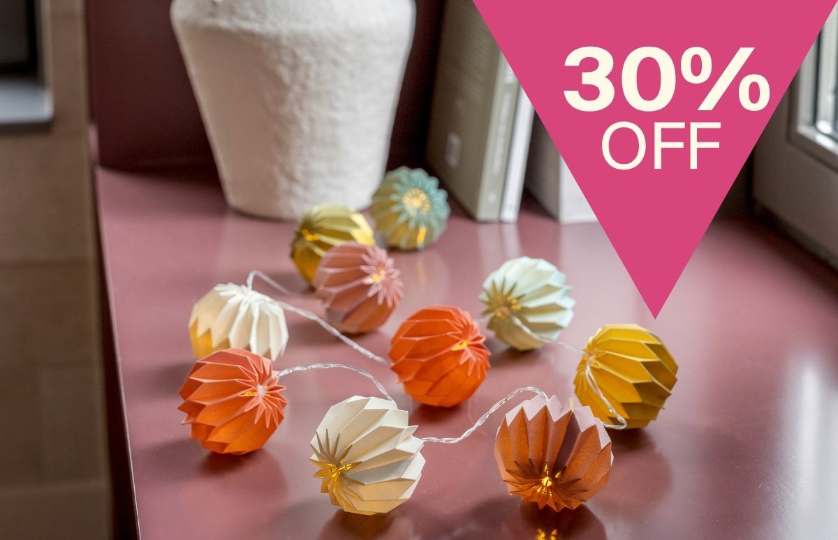 Eine Lichterkette mit zehn bunten, gefalteten Papierkugeln liegt auf einer rosafarbenen Oberfläche. Die Kugeln leuchten in warmen Tönen wie Orange, Gelb, Creme und Braun. Oben rechts: „30% OFF“.