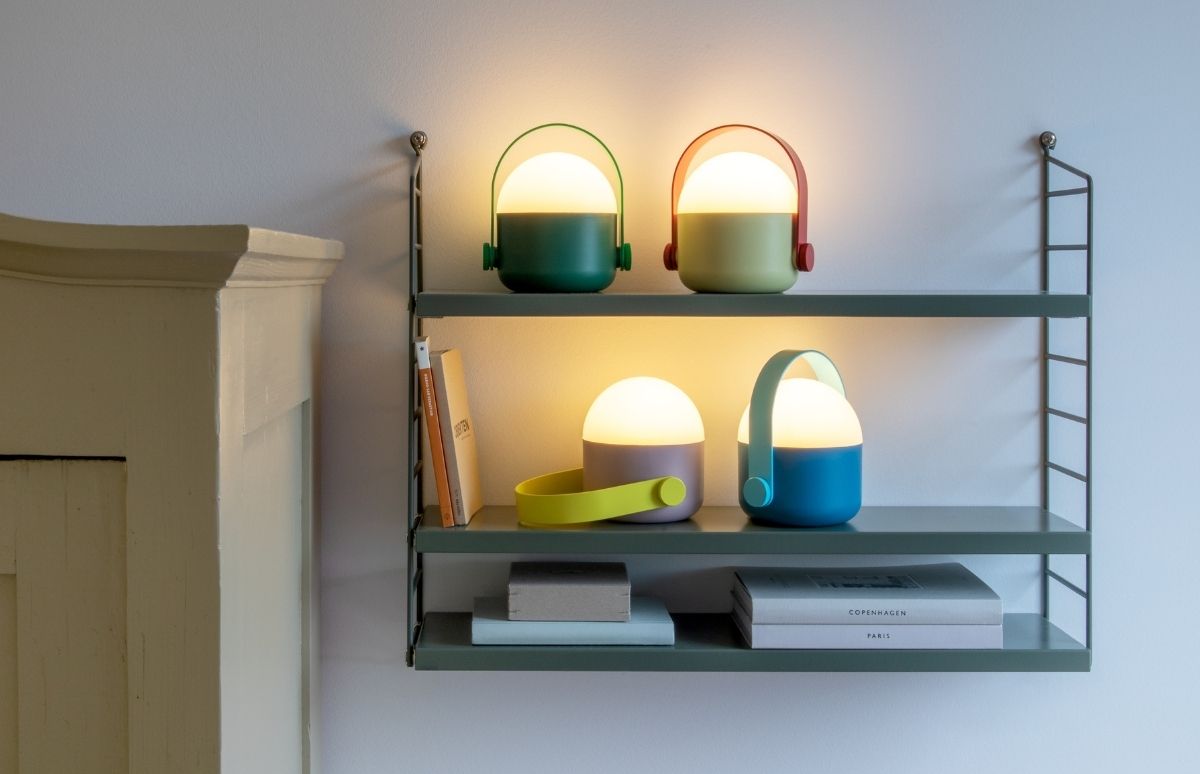 Vier portable LED-Lampen in Pastell- und Grüntönen stehen auf zwei dunkelgrünen Wandregalen. Oben: dunkelgrüne Lampe und hellgrüne mit rotem Griff. Unten: lila mit gelbem Band und blaue mit hellblauem Griff. Bücher daneben.