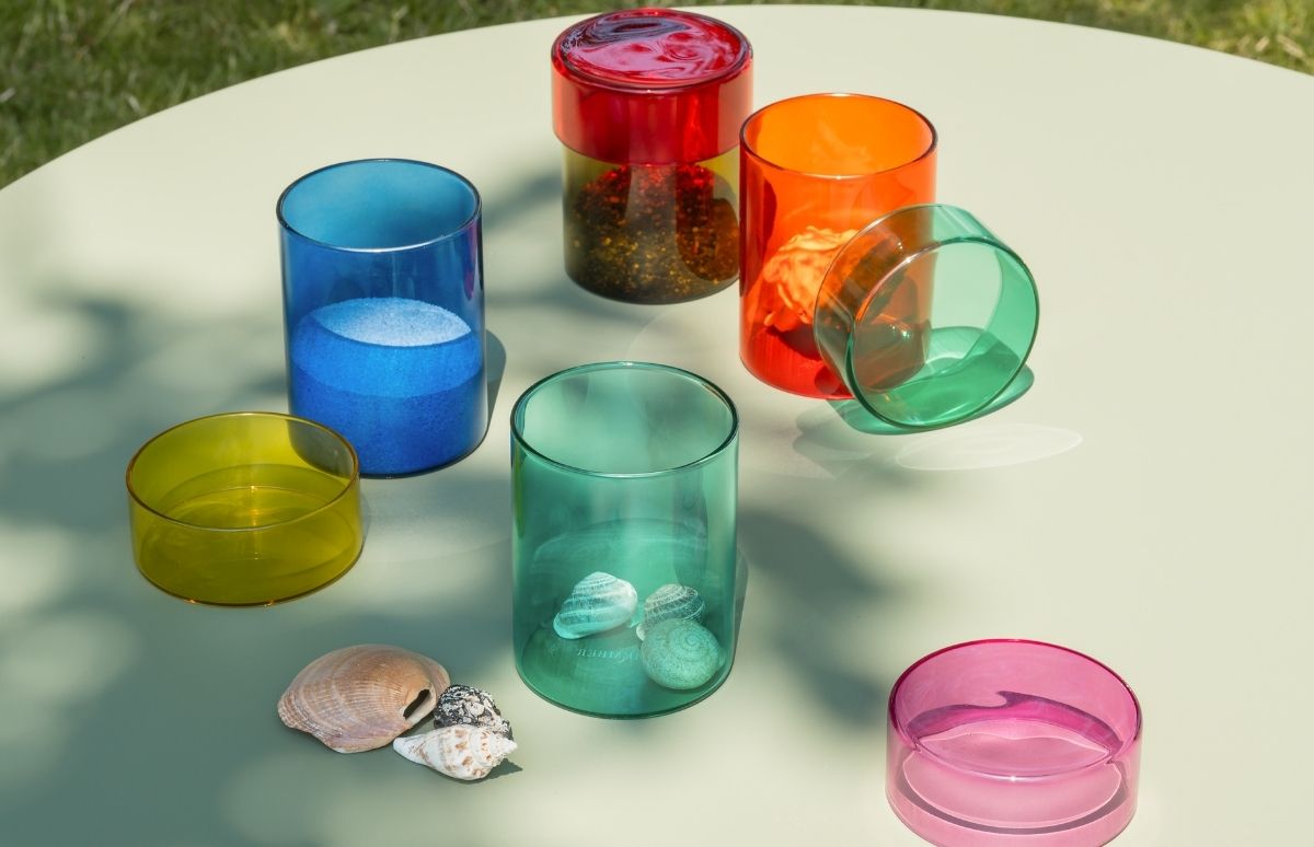 Sieben farbige Glasgefäße und Muscheln auf hellgrünem Tisch im Freien. Blaues Glas mit Sand, rotes mit dunklen Körnern, oranges mit getrockneter Blume, türkises mit kleinen Muscheln. Mehr Muscheln. Gelb und pink leer. Sonne wirft Schatten.