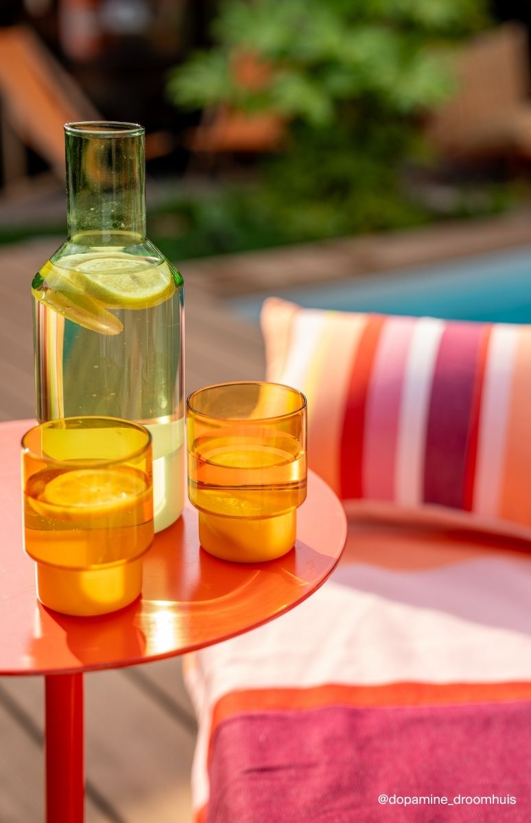 Erfrischende Szene am Pool: Auf einem roten Rundtisch stehen eine grüne Glasflasche mit Wasser und Limettenscheiben sowie zwei gelb-orangefarbene Gläser mit Orangenscheiben. Im Hintergrund sind Kissen und blauer Pool zu sehen.