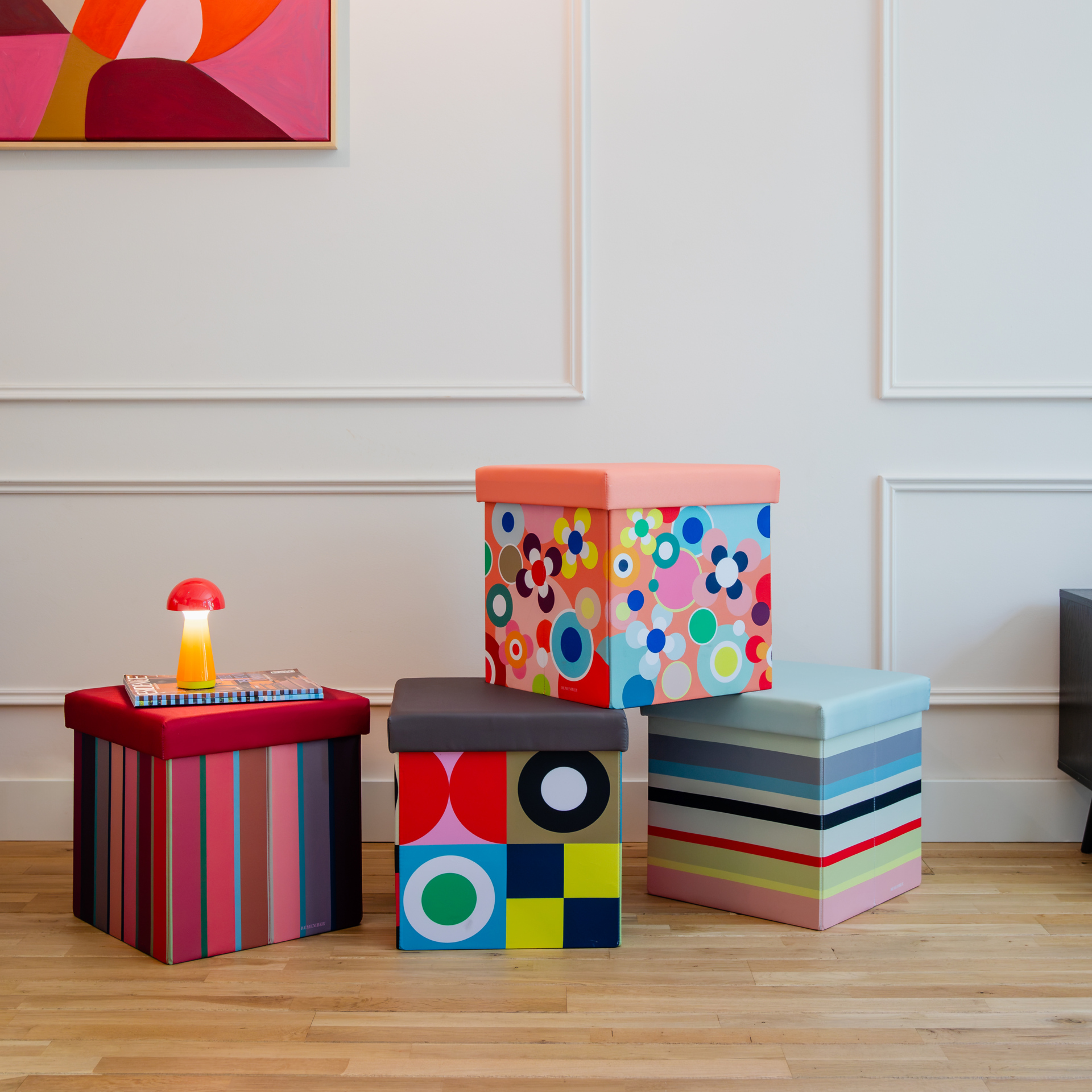 Vier bunte Remember Sitzboxen Costa Rossa mit Mustern – Streifen, geometrische Formen und florale Punkte – stehen auf hellem Holzboden. Teils gestapelt dienen sie als farbenfrohe, dekorative Aufbewahrungslösung.