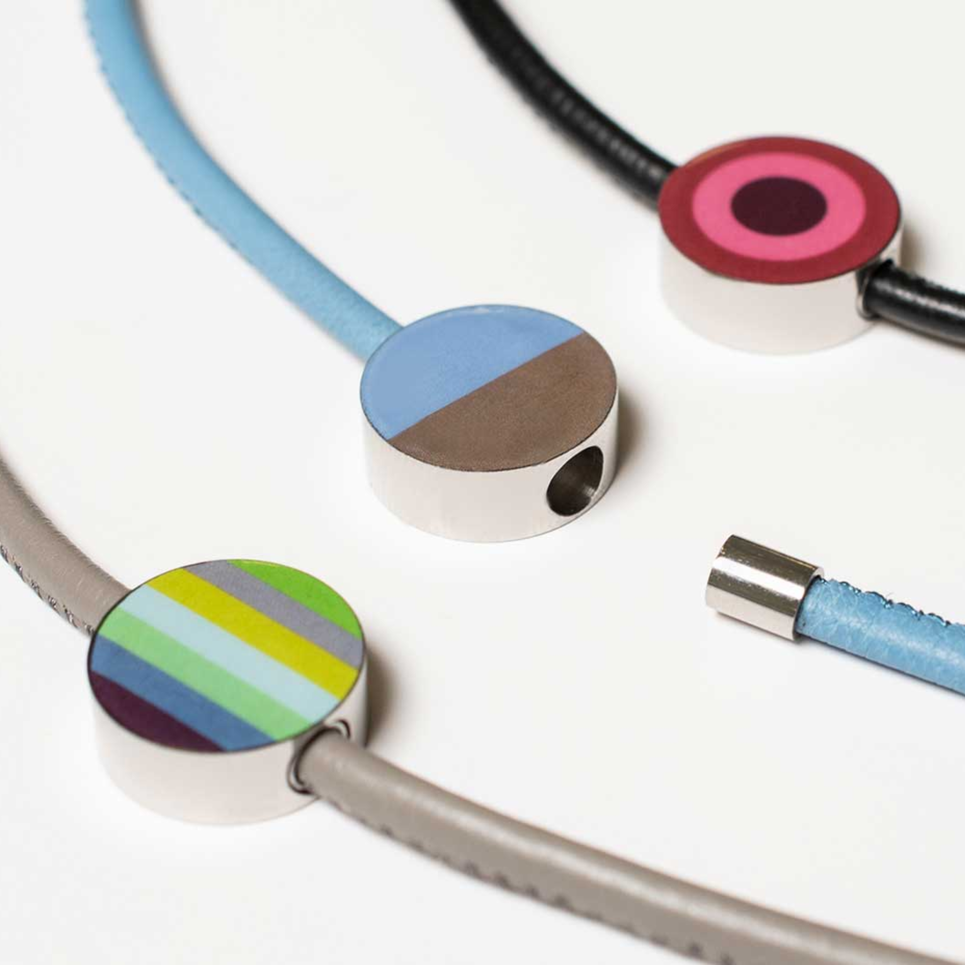 Gros plan sur trois pendentifs bijoux attachés à des rubans colorés. Les pendentifs ronds en acier inoxydable sont de couleurs différentes : rayés, bicolores et cercles concentriques. Un ruban est détaché du pendentif.