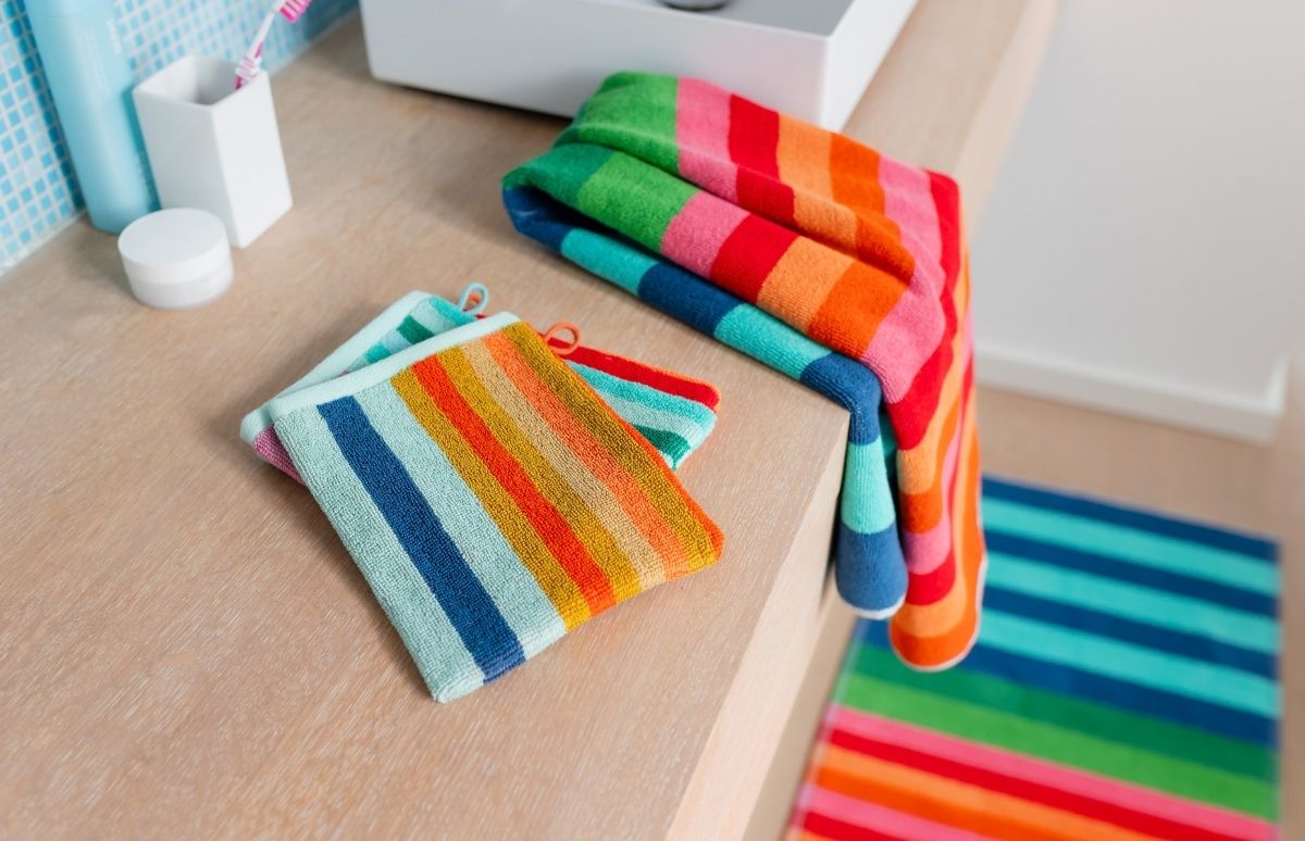 Zwei gefaltete gestreifte Waschlappen und ein größeres Handtuch liegen auf einer hellen Holztheke im Bad. Leuchtende Querstreifen in Blau, Grün, Gelb, Orange und Rot. Im Hintergrund blaue Fliesen, ein Zahnbürstenhalter und ein Badteppich.